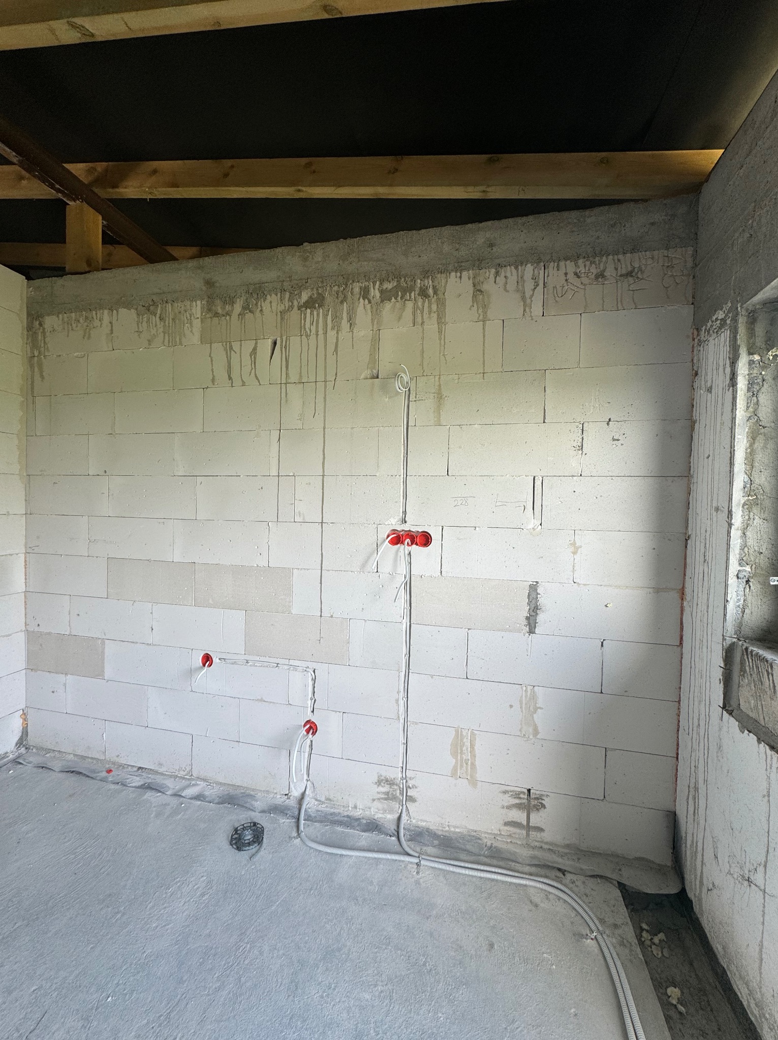 Surowy stan wnętrza w trakcie prac: ściany z białych bloczków, widoczne przewody elektryczne z czerwonymi puszkami podtynkowymi, instalacja podłogowa w peszlach, betonowa wylewka, drewniana...