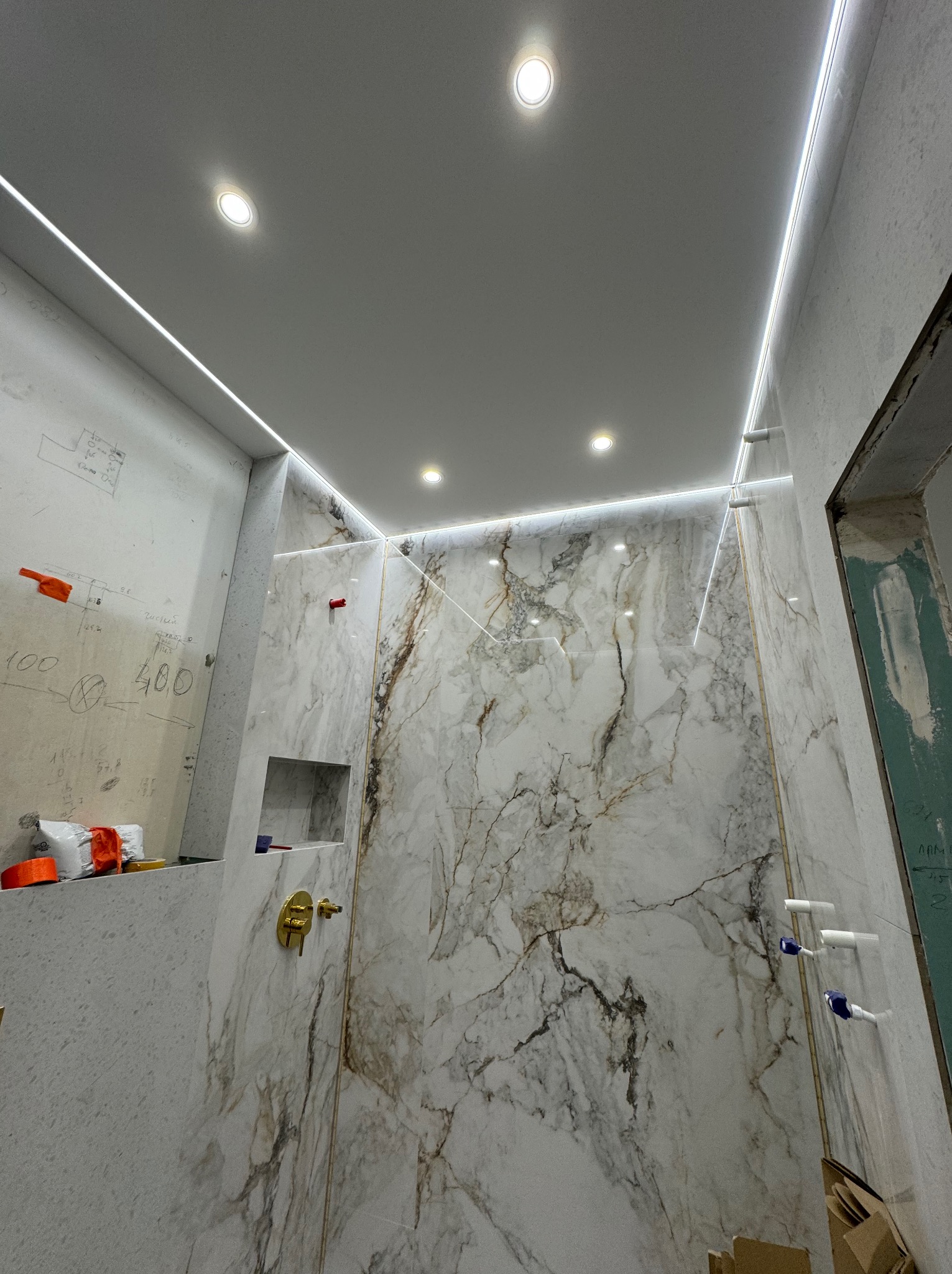 Wnętrze łazienki w trakcie wykańczania, widoczne marmurowe płytki na ścianach, złota armatura, oświetlenie LED na suficie oraz fragment ściany z naniesionymi wymiarami i notatkami.