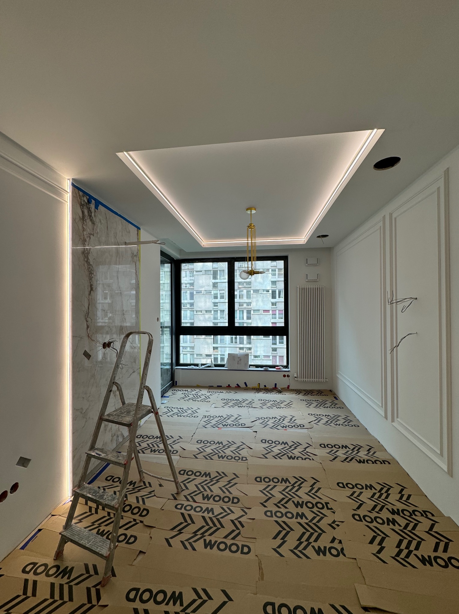 Pokój w trakcie wykańczania z zabezpieczoną podłogą, marmurową ścianą z podświetleniem LED, dekoracyjnymi listwami ściennymi, nowoczesną lampą i metalową drabiną.