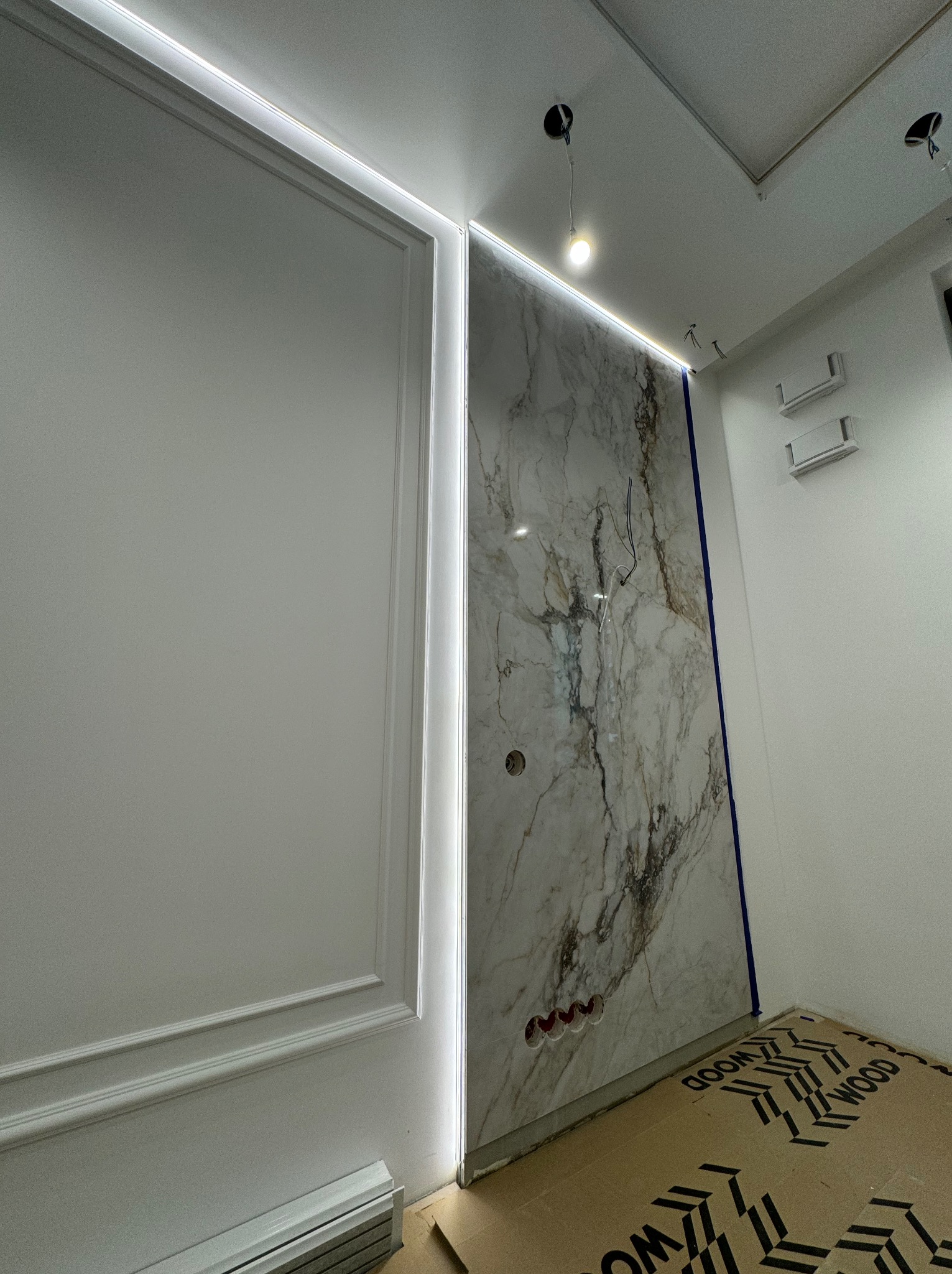 Ściana z marmurowym panelem podświetlonym listwą LED, obok ściana z dekoracyjną sztukaterią, widoczne przygotowane otwory na gniazdka elektryczne i wystające przewody.