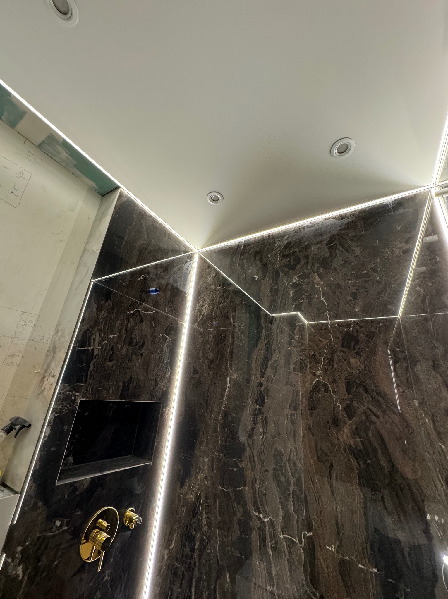 Łazienka w trakcie wykańczania: ciemny marmur z podświetleniem LED, złota bateria podtynkowa, wnęka w ścianie, sufit z oświetleniem punktowym.