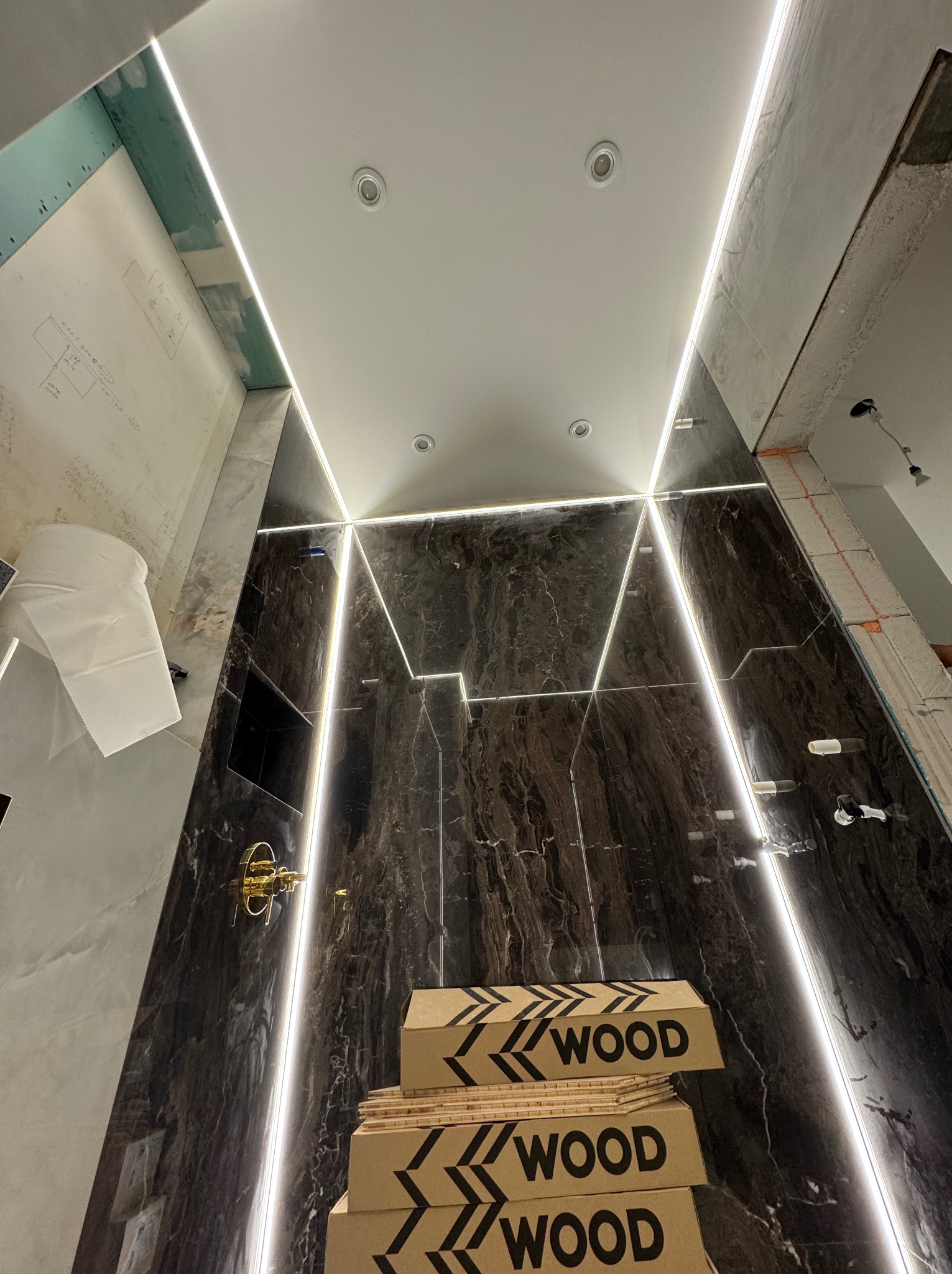 Łazienka w trakcie wykończenia, widoczne marmurowe płytki, oświetlenie LED w kształcie trójkątów, złota armatura, kartony z napisem WOOD i fragmenty instalacji elektrycznej na ścianie.