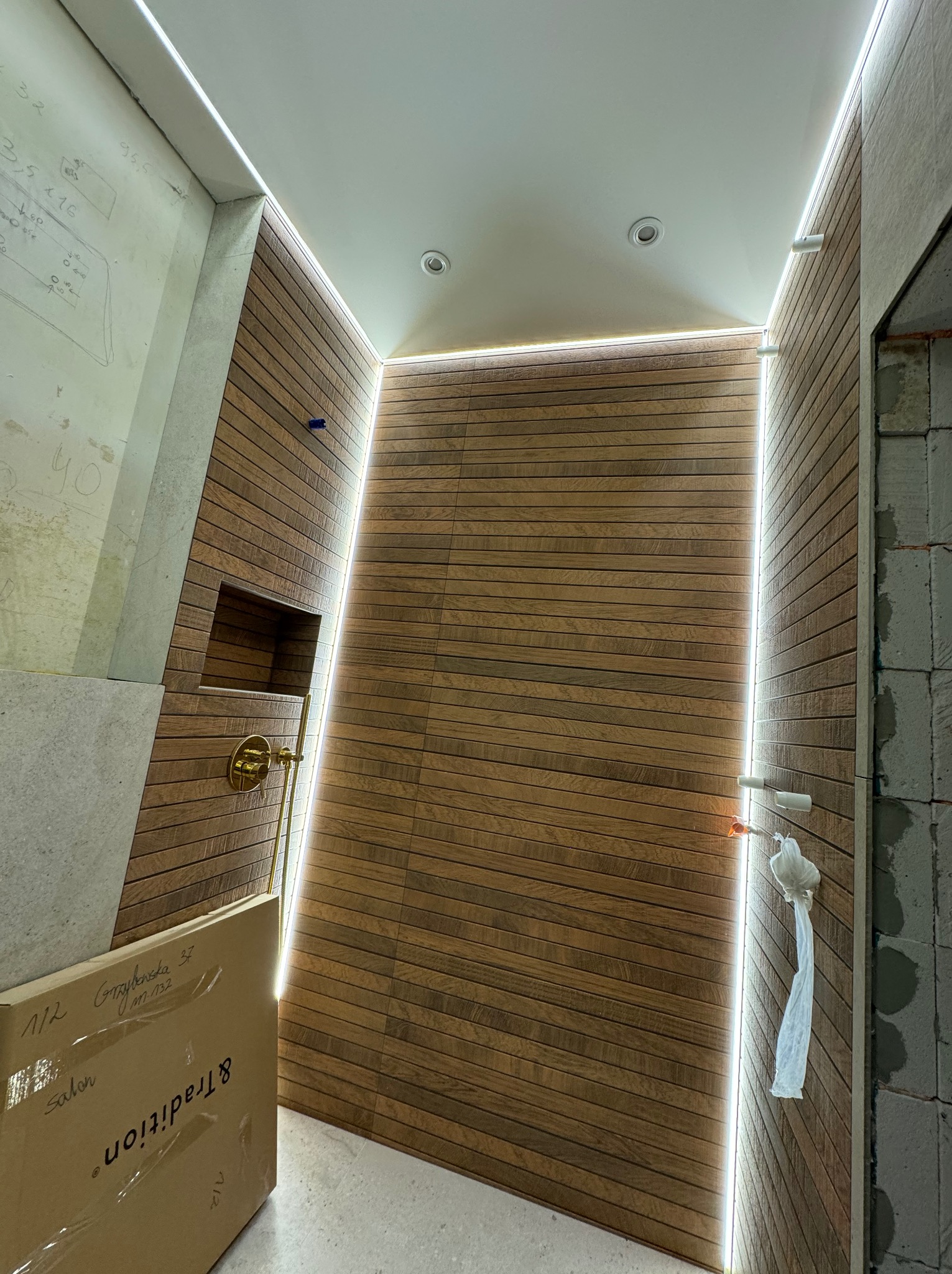 Nowoczesna łazienka w trakcie wykańczania, z drewnopodobnymi płytkami ułożonymi poziomo, podświetleniem LED wzdłuż krawędzi, złotą armaturą w niszy i kartonem z napisem 'Tradition' w rogu.