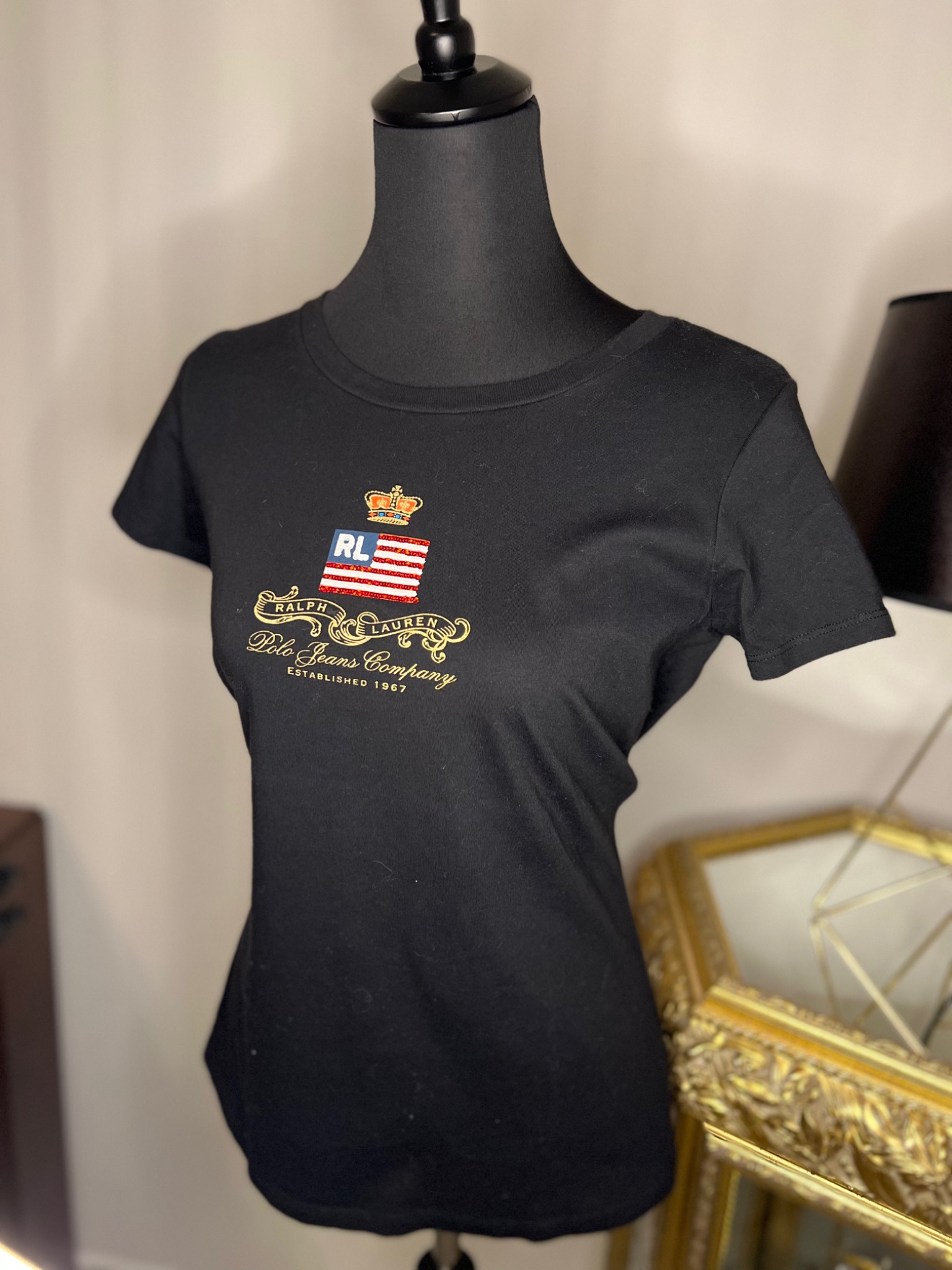 Czarna koszulka damska z krótkim rękawem, z haftowanym logo Polo Jeans Company przedstawiającym koronę i flagę, prezentowana na czarnym manekinie na tle beżowej ściany i fragmentu złotej ramy.