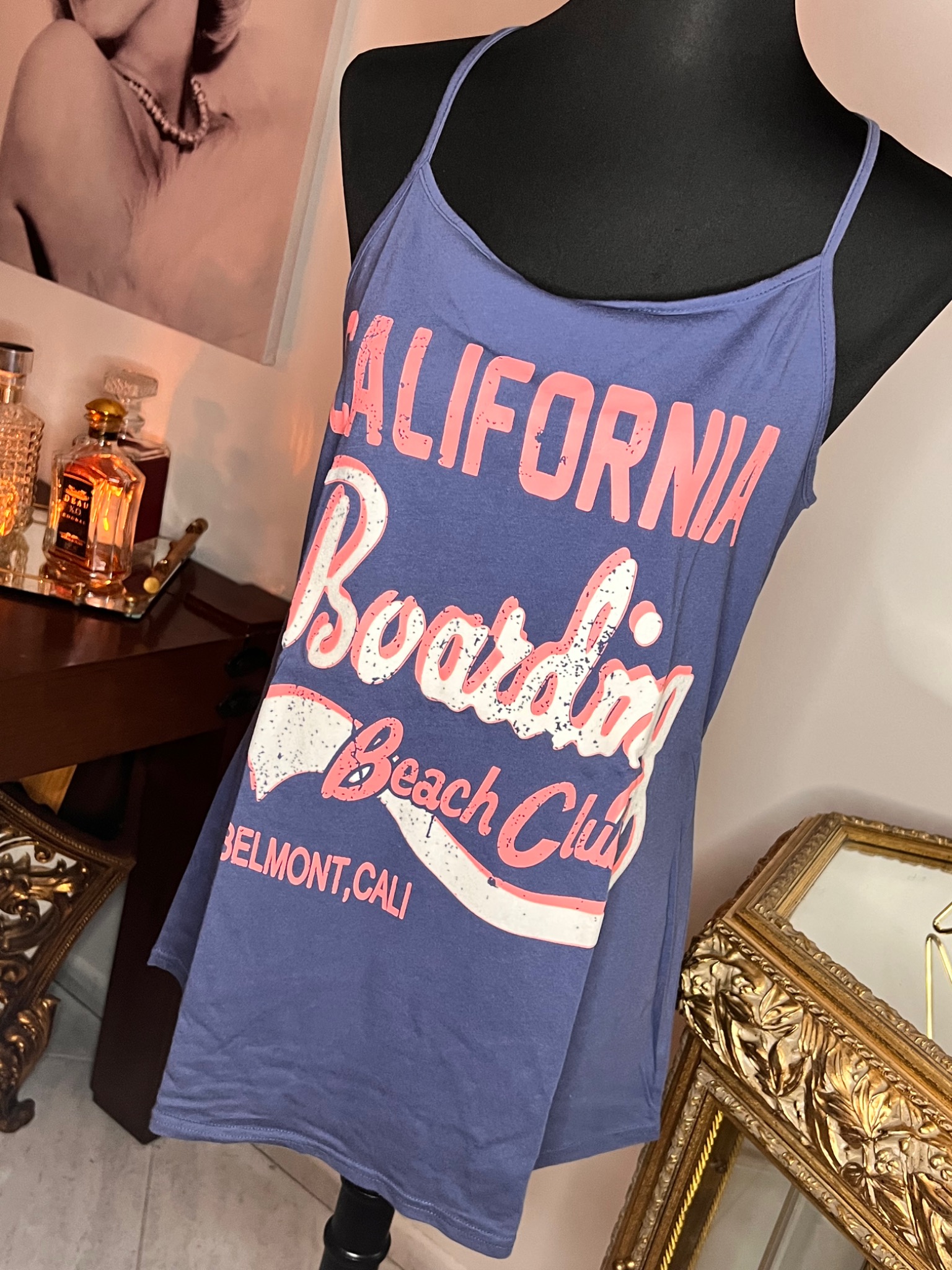 Niebieska koszulka na ramiączkach z nadrukiem 'California Boarding Beach Club' na manekinie, obok złotej ramy lustra i flakonów perfum