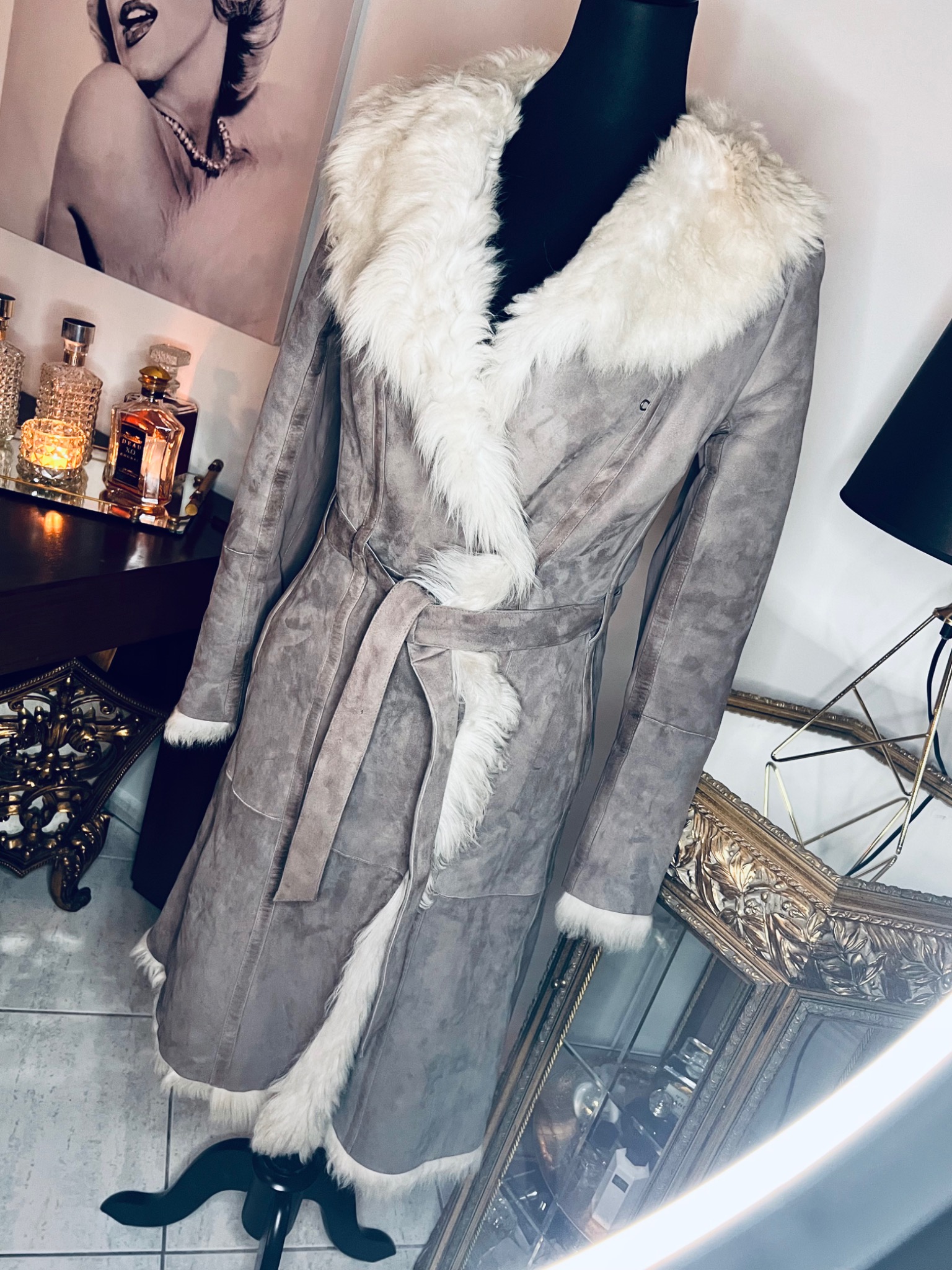Długi szary płaszcz z futrzanym kołnierzem i wykończeniami, zapinany na pasek, prezentowany na manekinie na tle lustra w ozdobnej złotej ramie, flakonów perfum i portretu Marilyn Monroe.