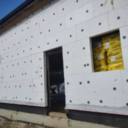 Ocieplenie budynku styropianem z widocznymi czarnymi kołkami mocującymi, przygotowane otwory okienne i drzwiowe, żółte paczki styropianu w otworze okiennym.