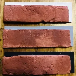 Trzy próbki cegieł klinkierowych w odcieniach czerwieni, każda na podkładzie w innym kolorze: białym, szarym i czarnym, ułożone na drewnianym blacie.