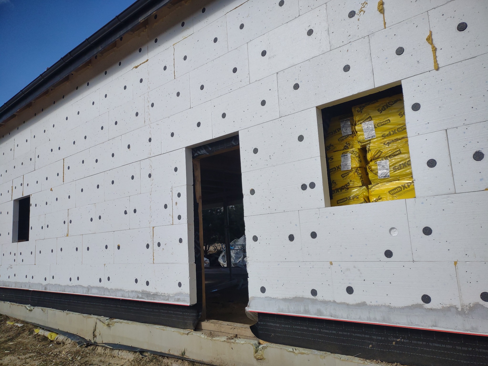 Ocieplenie budynku styropianem z widocznymi czarnymi kołkami mocującymi, przygotowane otwory okienne i drzwiowe, żółte paczki styropianu w otworze okiennym.