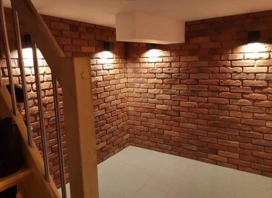 Wykończona ściana z cegły dekoracyjnej w pomieszczeniu z drewnianymi schodami i nowoczesnym oświetleniem punktowym.