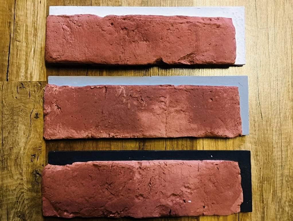 Trzy próbki cegieł klinkierowych w odcieniach czerwieni, każda na podkładzie w innym kolorze: białym, szarym i czarnym, ułożone na drewnianym blacie.