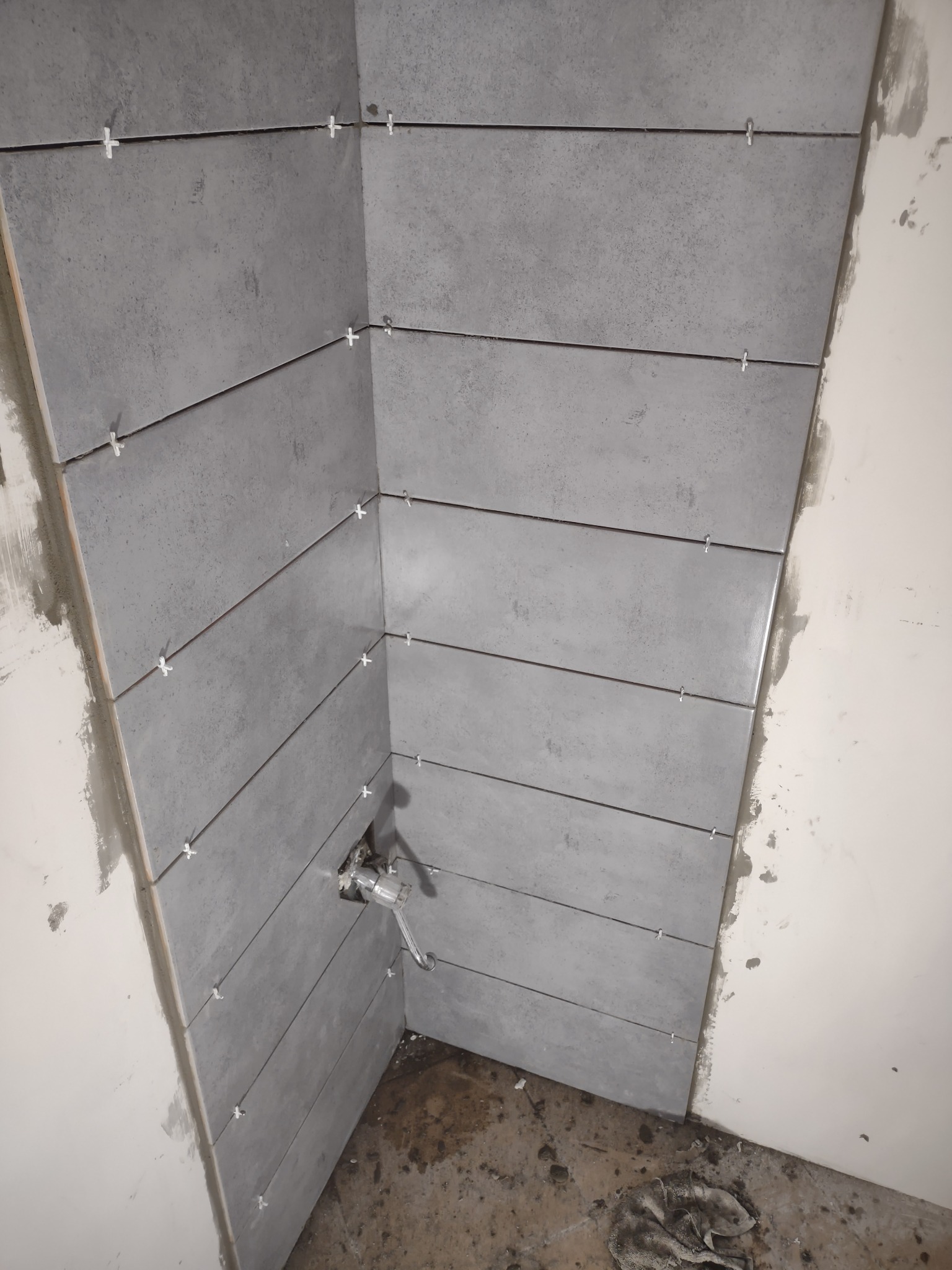 Szare, prostokątne płytki ceramiczne układane poziomo na ścianie w rogu łazienki, z krzyżykami dystansowymi, widoczny fragment instalacji wodnej i niedokończone wykończenie ściany.