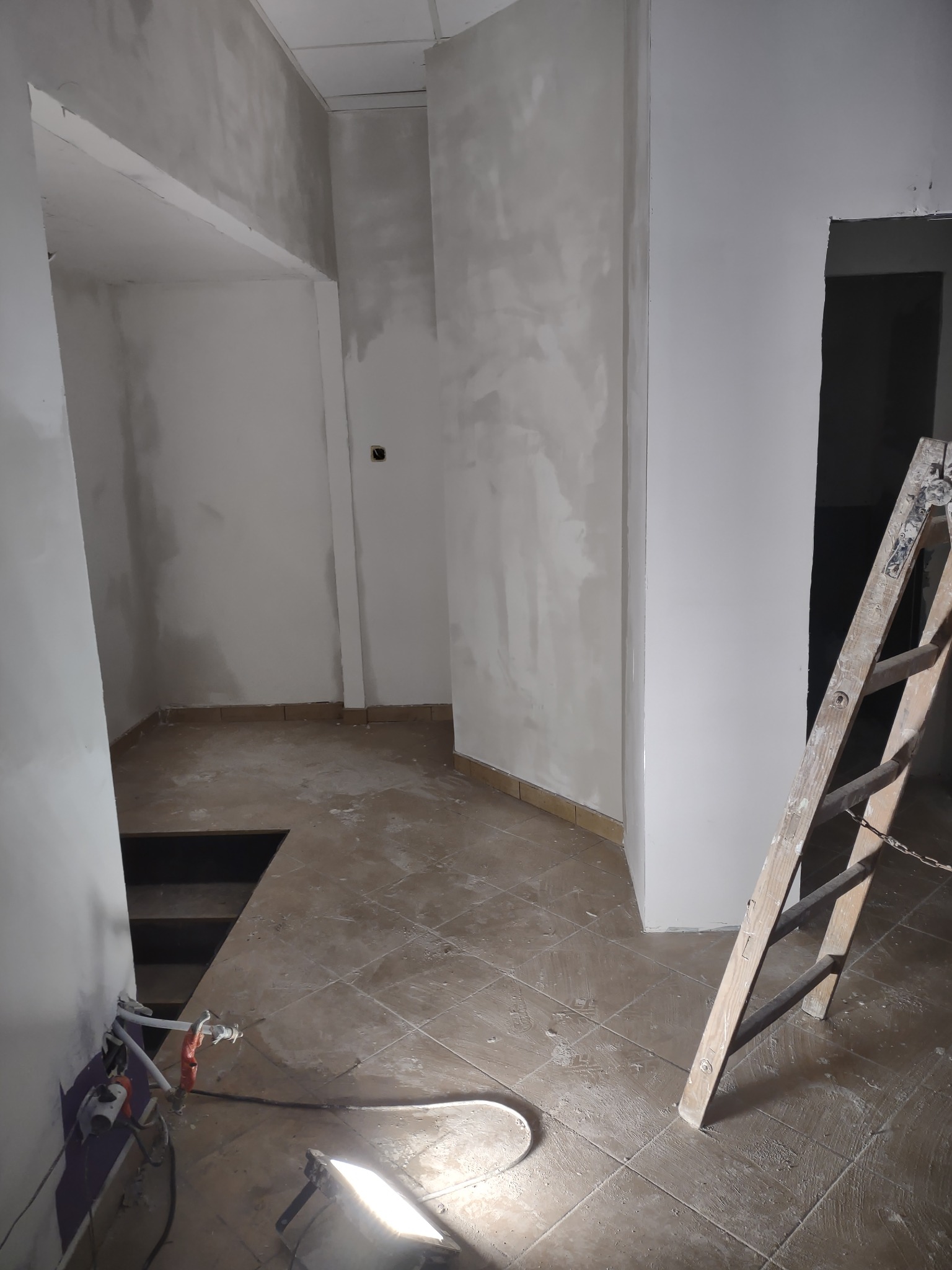 Wnętrze w trakcie prac wykończeniowych: ściany pokryte gładzią, widoczna drabina, fragment instalacji elektrycznej i otwór w podłodze.