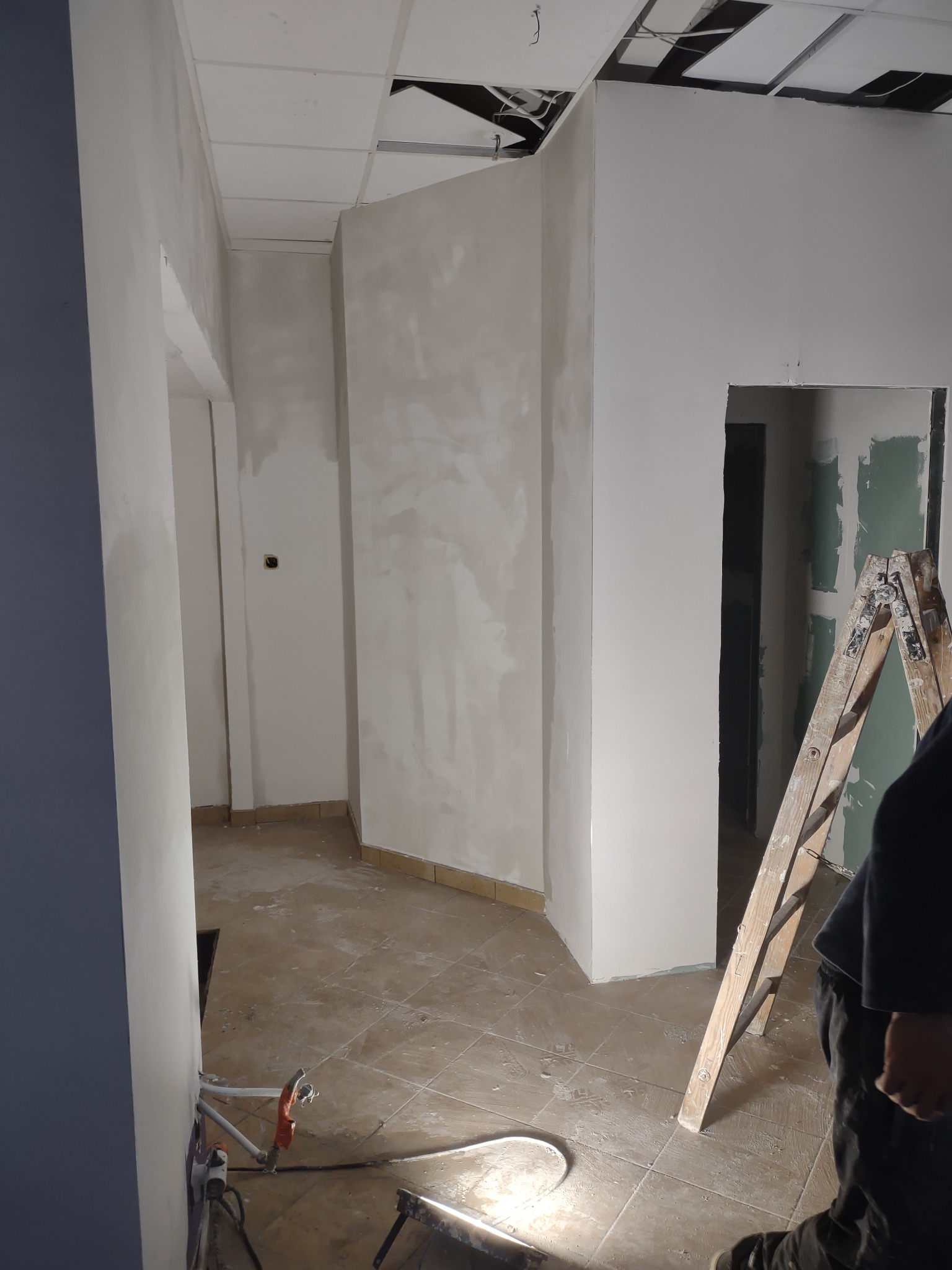 Wnętrze w trakcie prac wykończeniowych: ściany po gładzi, częściowo podwieszony sufit z widocznymi instalacjami, płytki na podłodze, drabina oparta o ścianę, widoczne narzędzia i sprzęt.