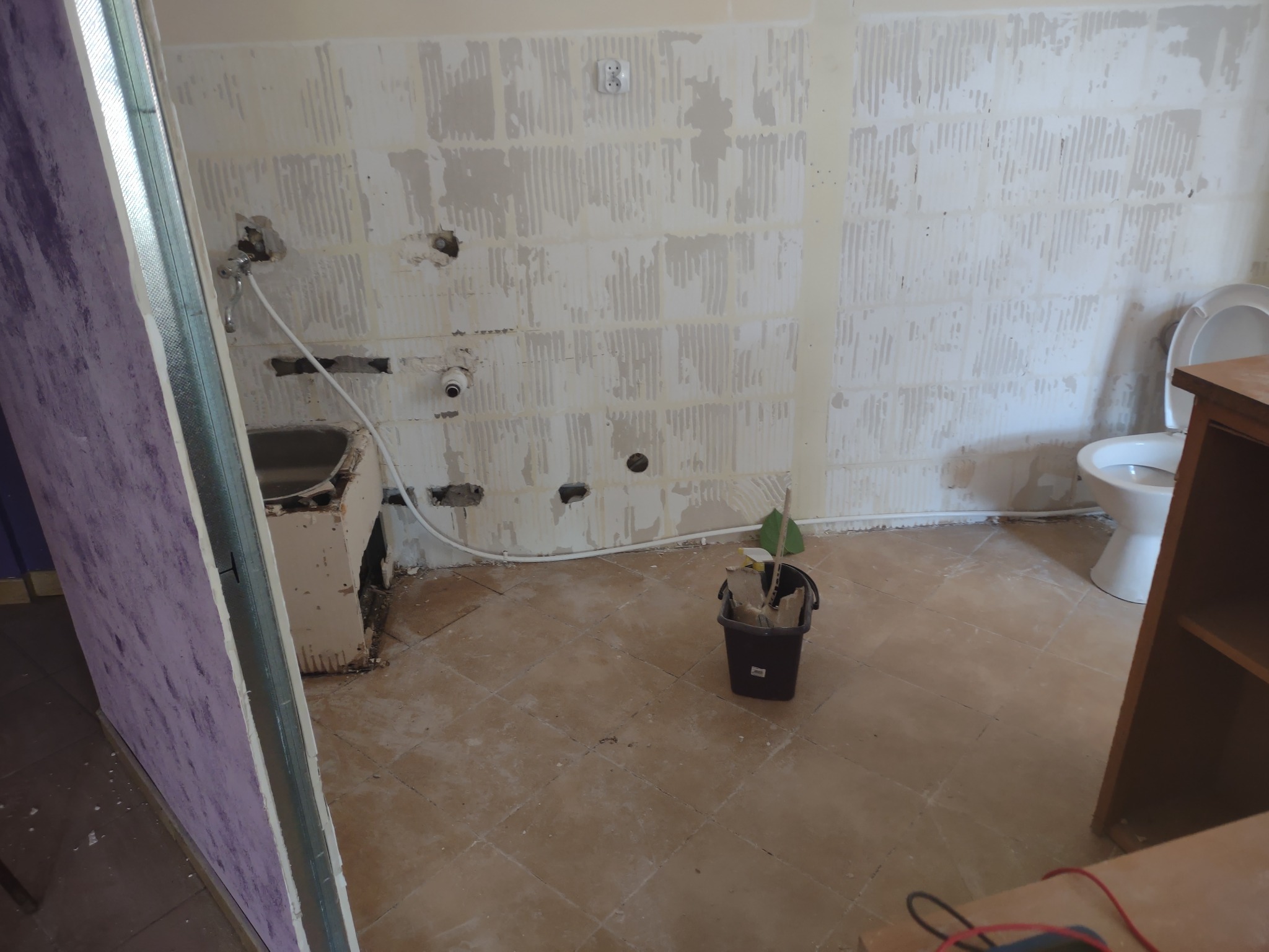 Łazienka w trakcie demontażu płytek, widoczne ślady po kleju na ścianach, przygotowania do remontu, częściowo zasłonięta toaleta, kubeł z narzędziami na podłodze.