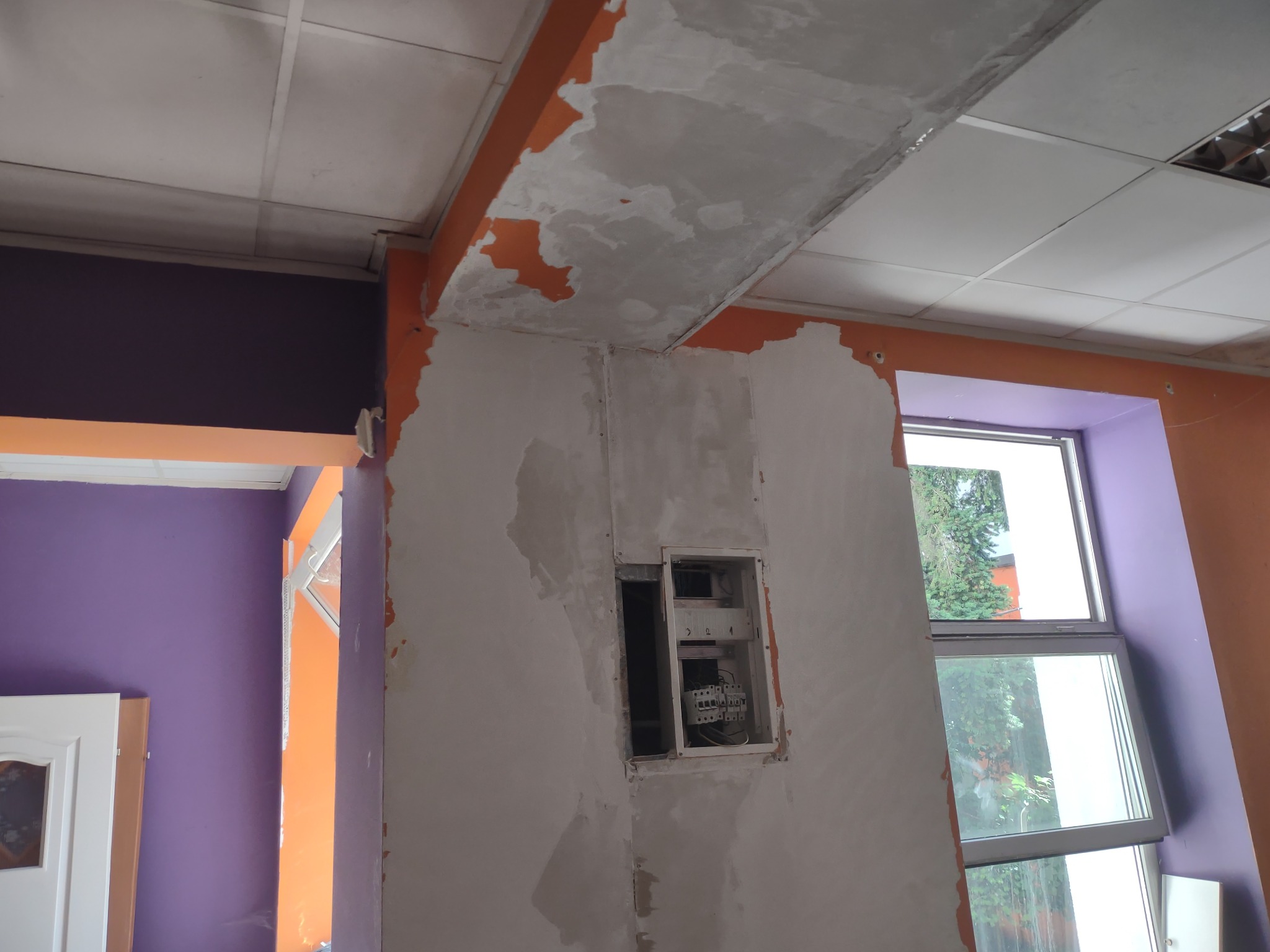 Ściana w trakcie prac wykończeniowych z otwartą skrzynką elektryczną, widoczne przewody i bezpieczniki, ściany częściowo pomalowane na pomarańczowo i fioletowo, sufit kasetonowy.
