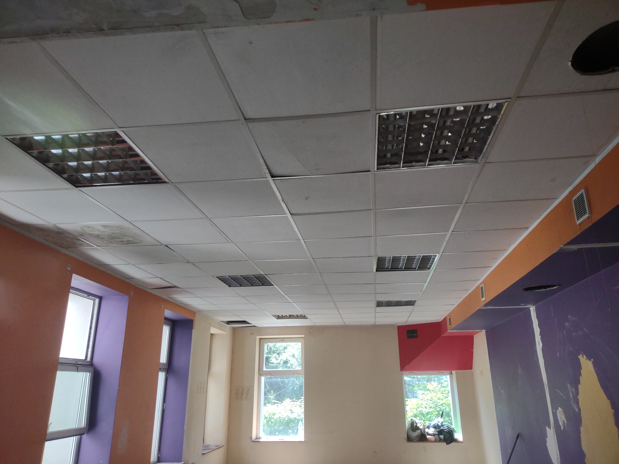 Sufit podwieszany kasetonowy w trakcie montażu, widoczne otwory na oświetlenie i niedokończone ściany w pomieszczeniu z oknami.