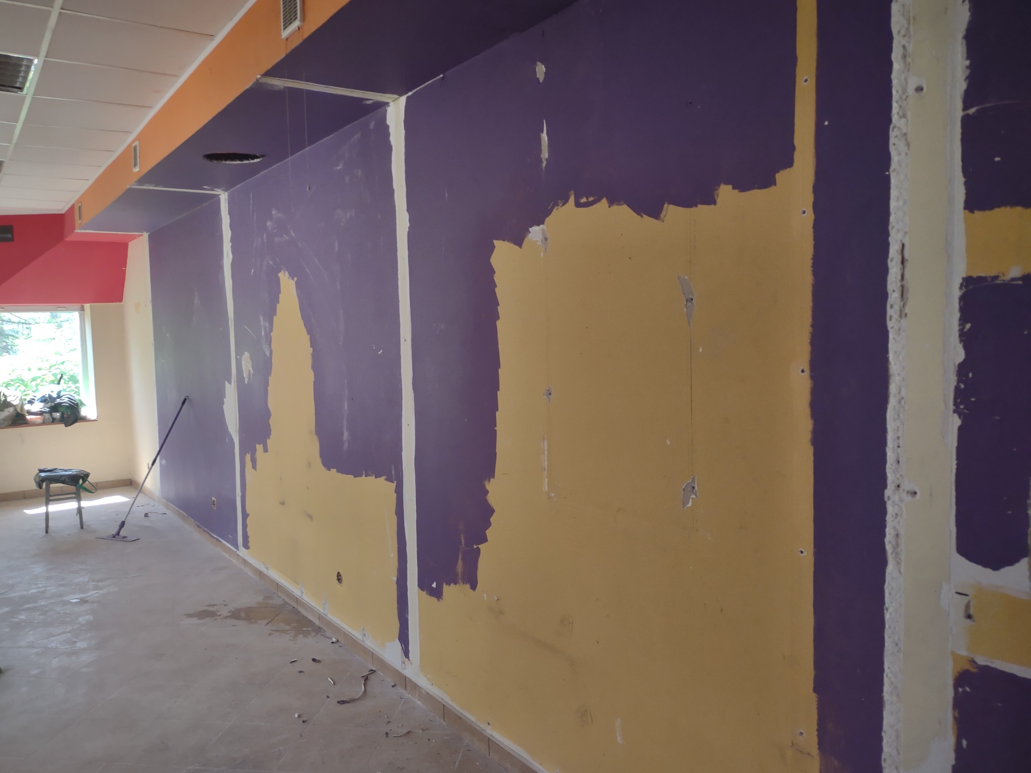 Ściana w trakcie malowania na fioletowo, widoczne fragmenty żółtej farby, niedokładne wykończenie, mop na podłodze, widoczne ślady po remoncie, dziury w ścianie.