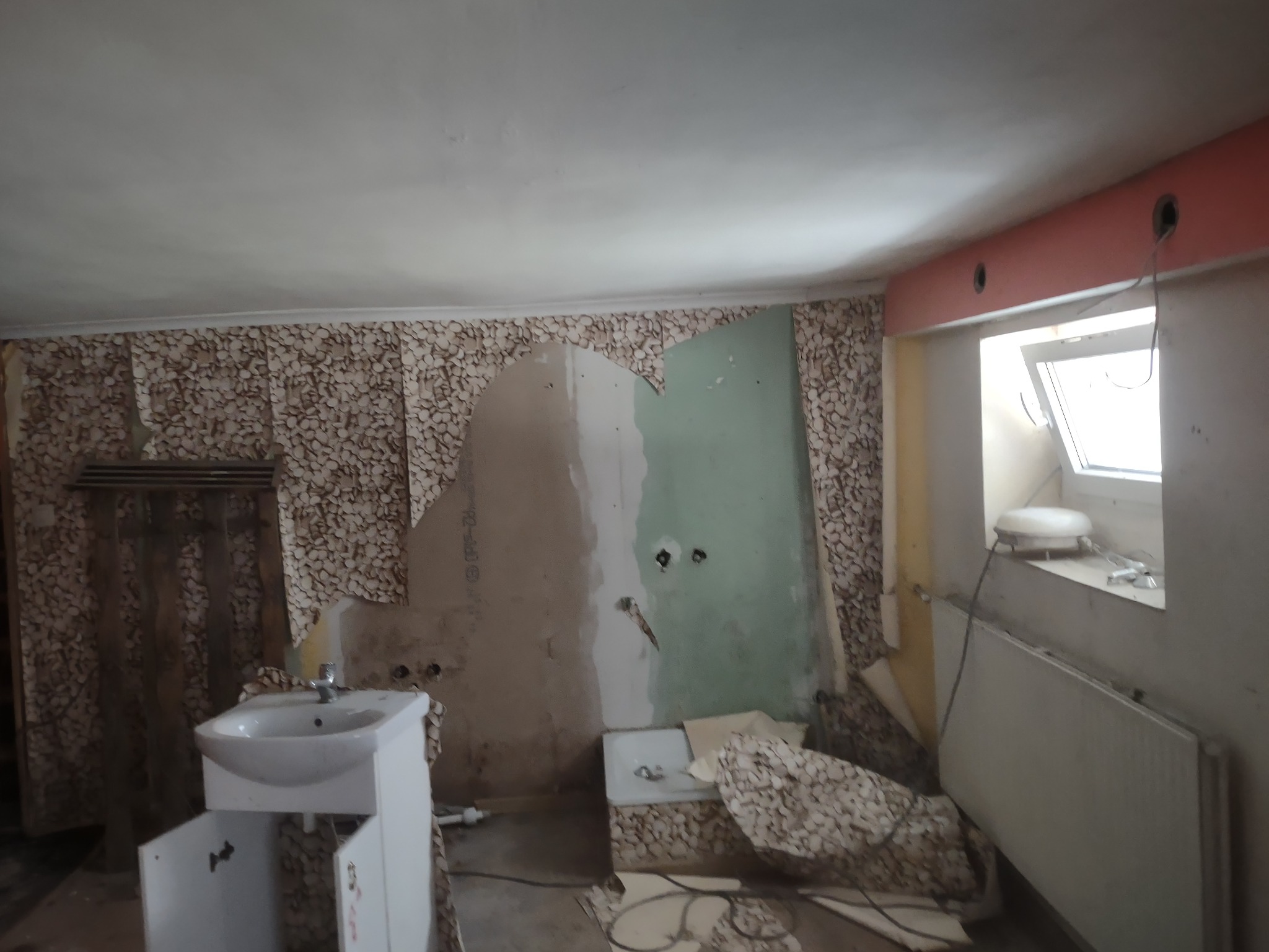 Demontaż starej tapety w łazience podczas remontu, widoczny fragment wanny, umywalka z szafką i grzejnik, ściany częściowo przygotowane do dalszych prac wykończeniowych.