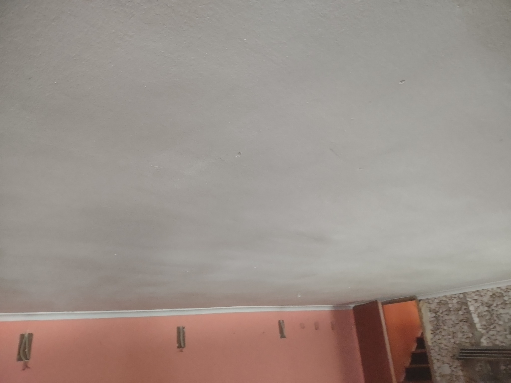 Świeżo pomalowany sufit w pokoju, widoczne niedoskonałości wykończenia, ściany w kolorze brzoskwiniowym z wystającymi puszkami elektrycznymi, fragment ściany pokryty tapetą strukturalną, otwarte...