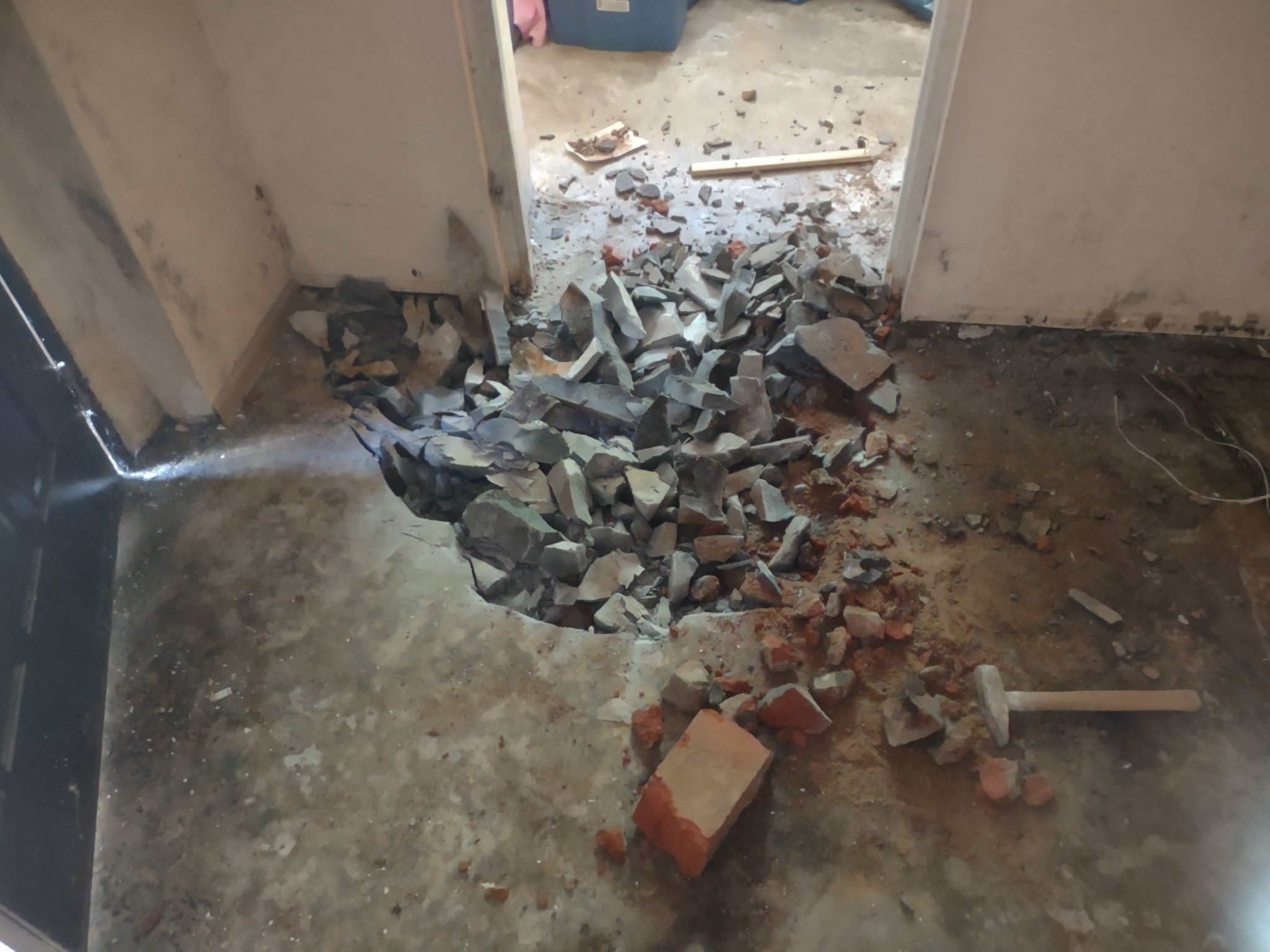 Demontaż podłogi: fragment wyburzonej posadzki z gruzem i cegłami, widoczny młotek i fragment listwy przypodłogowej w przejściu do kolejnego pomieszczenia.