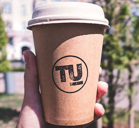 Ręka trzyma papierowy kubek z logo 'TU' na tle rozmytej zieleni i budynku.