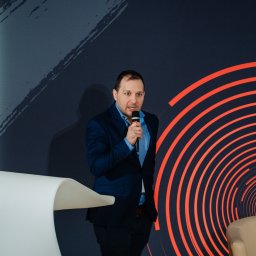 Dotono - Mężczyzna w garniturze przemawia do mikrofonu na tle ściany z abstrakcyjnym wzorem. Białe krzesło i fragment fotela w kadrze.