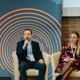 Dotono - Konferencja biznesowa: mężczyzna w garniturze przemawia do mikrofonu, obok siedzi kobieta w kwiecistej sukience. W tle grafika z pomarańczowymi okręgami.
