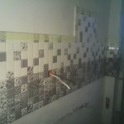 Układanie mozaiki z dekoracyjnych płytek w łazience, widoczne wystające kable elektryczne ze ściany, etap prac wykończeniowych.