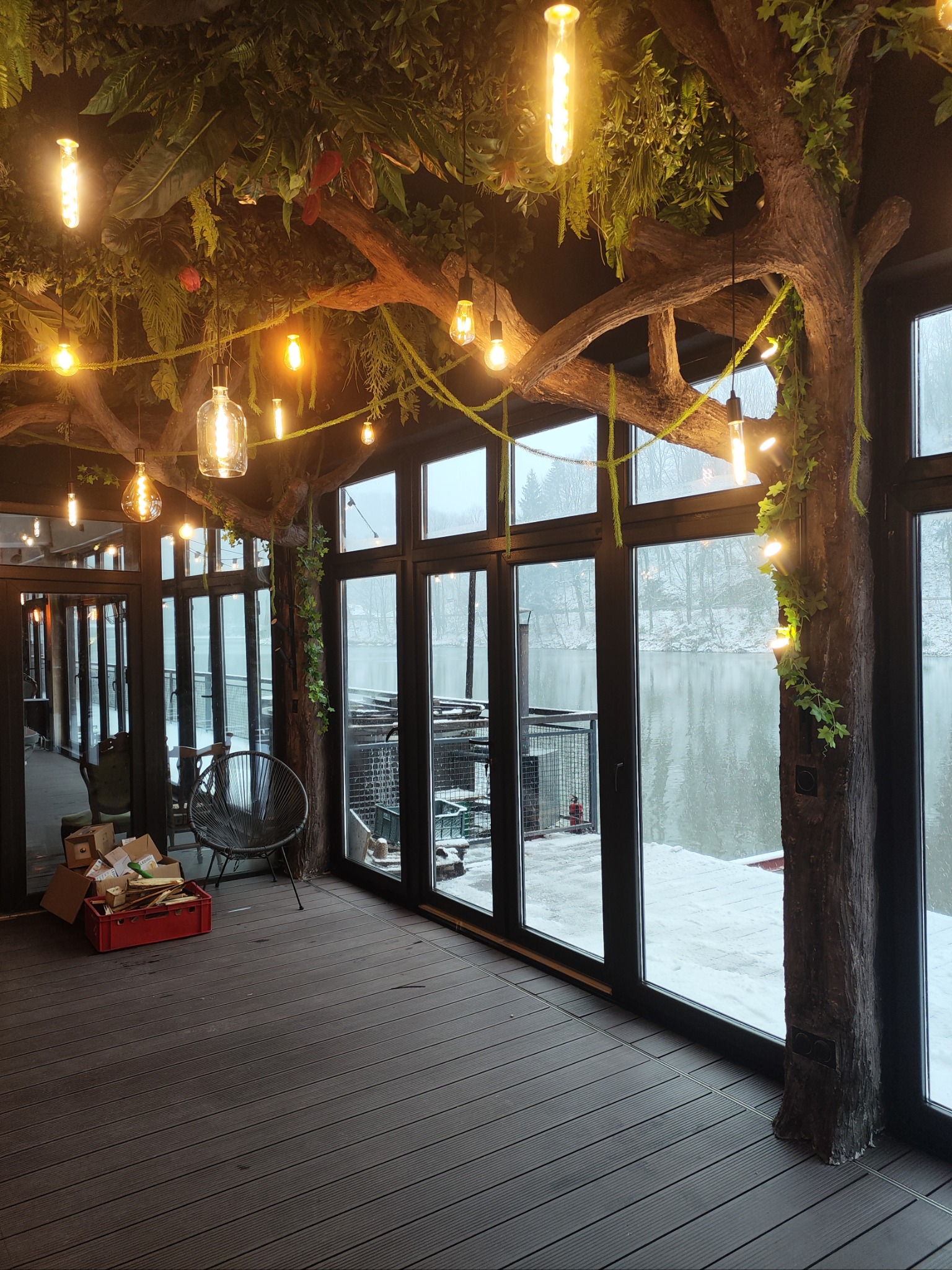 Wnętrze z drewnianą podłogą i dużymi oknami z widokiem na zaśnieżony krajobraz, sufit dekorowany sztuczną roślinnością i wiszącymi żarówkami, tworzące unikalny klimat.