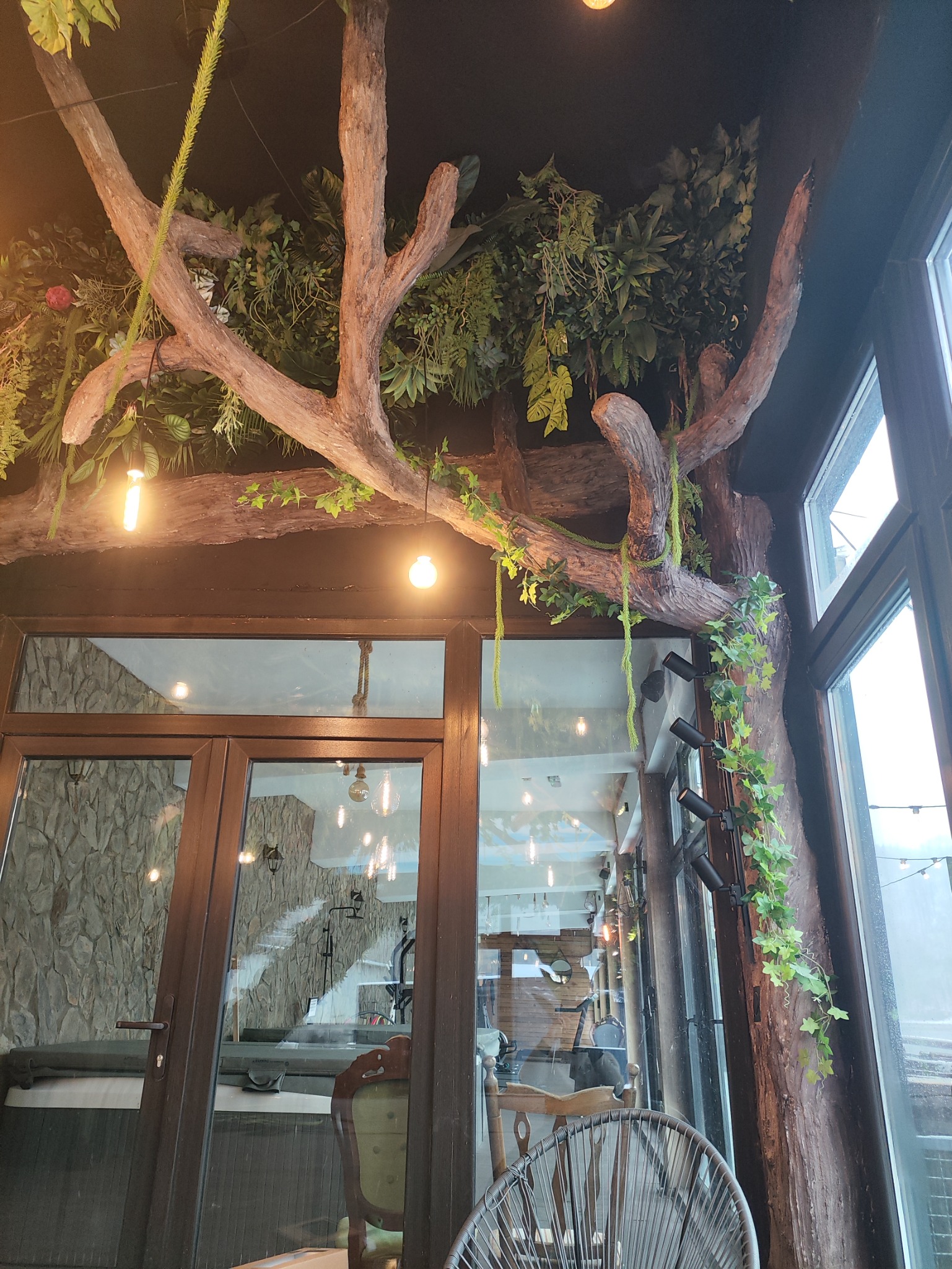 Dekoracyjny sufit z imitacją drzewa i roślinności w lokalu gastronomicznym, oświetlony wiszącymi żarówkami, widoczne okna i drzwi.