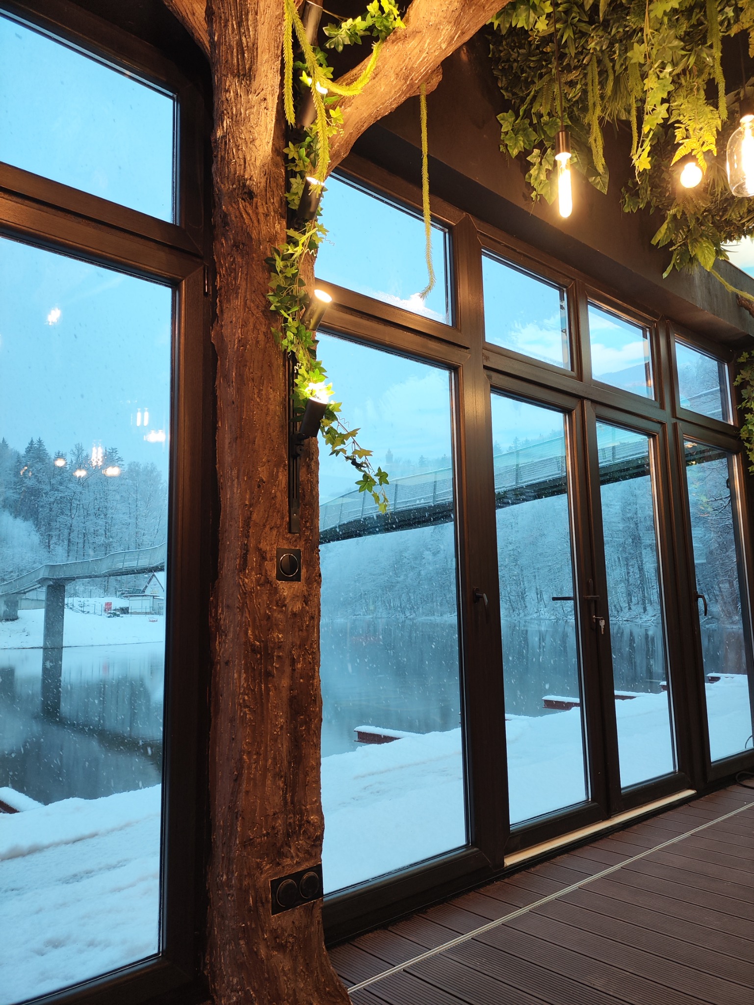 Wnętrze z dużymi oknami z widokiem na zimowy krajobraz z mostem, dekoracyjny pień drzewa z oświetleniem i roślinami na suficie.