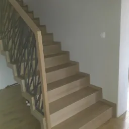 Jasne, drewniane stopnie z metalową, geometryczną balustradą na tle białej ściany z włącznikiem, widok z dołu.