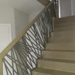 Drewniane schody z nowoczesną balustradą o geometrycznym wzorze, jasne drewno stopni i poręczy, szare ściany w tle.