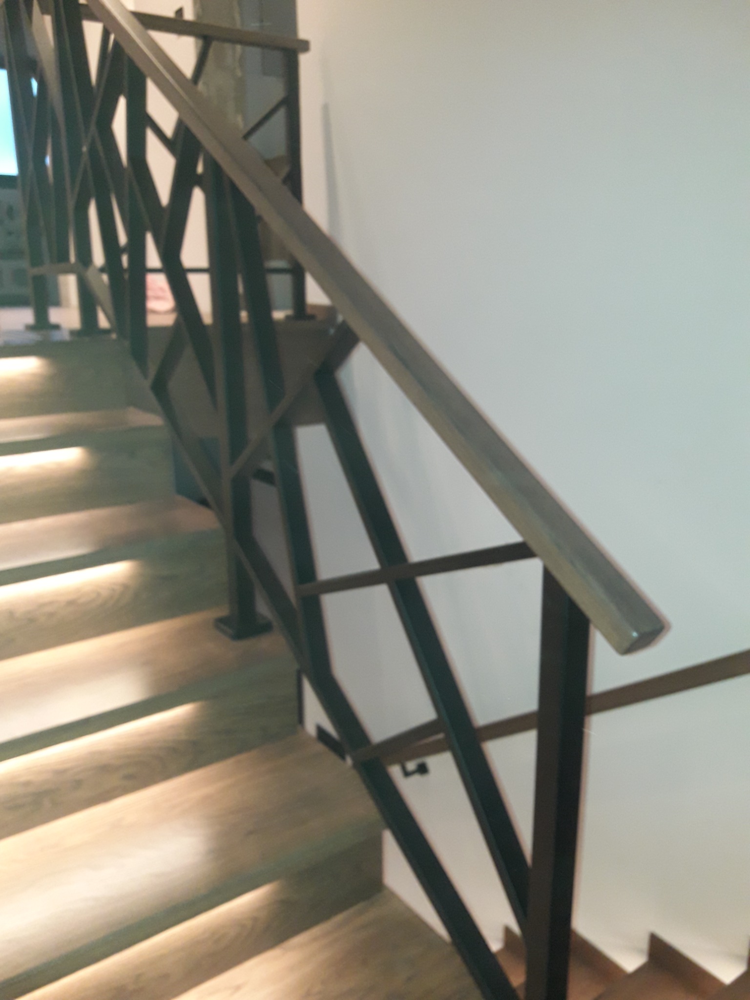 Nowoczesne schody z drewnianymi stopniami, podświetlane od spodu, i metalową, geometryczną balustradą w ciemnym kolorze.