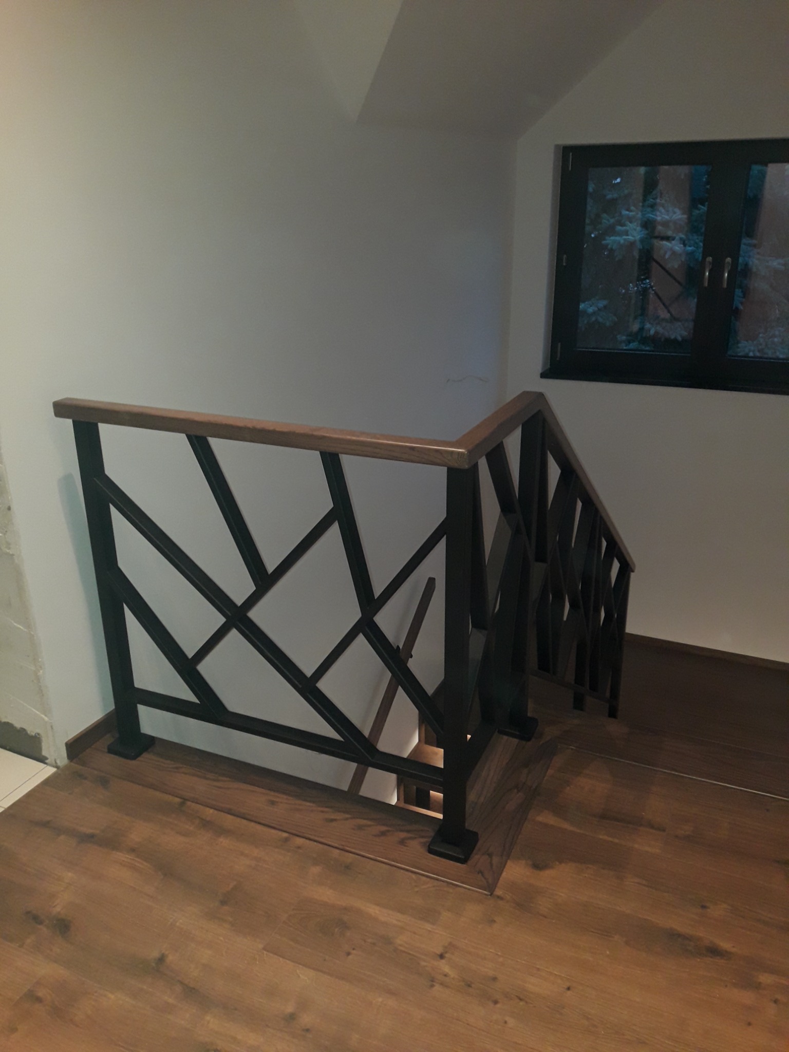 Nowoczesna balustrada schodowa z czarnych, geometrycznych elementów i drewnianą poręczą, widok z góry na drewniane stopnie i okno z widokiem na zieleń.