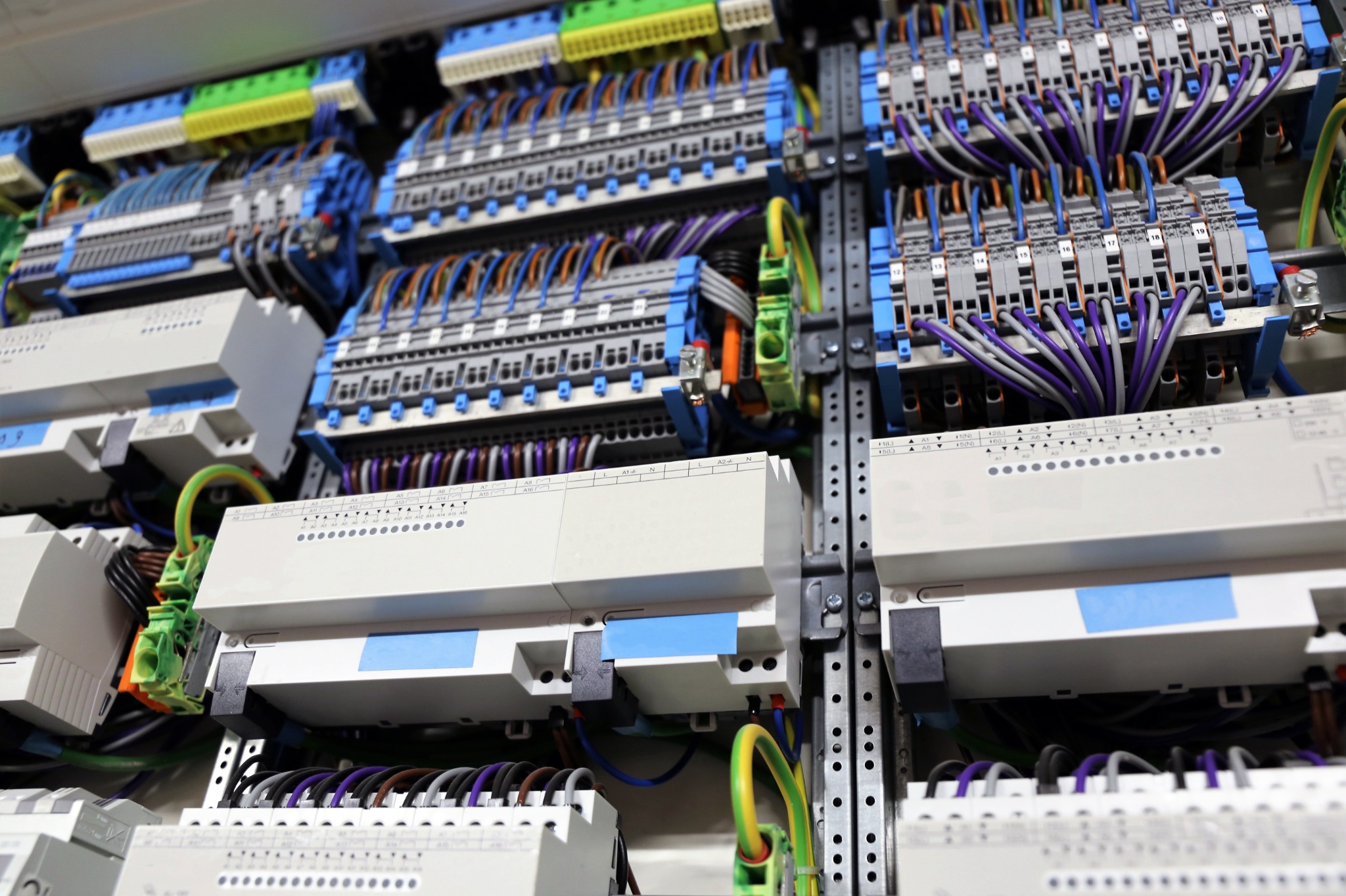 Rozbudowana szafa sterownicza z licznymi modułami, złączkami i okablowaniem w kolorach szarym, fioletowym, zielonym i pomarańczowym, zamontowana na perforowanej szynie.