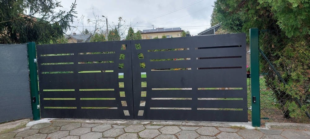 Nowoczesna, czarna brama wjazdowa z poziomymi i kwadratowymi wycięciami, zamontowana na zielonych słupkach, widok z zewnątrz na tle brukowanej nawierzchni i zieleni.
