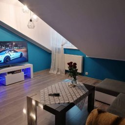 Dawid Mielewczyk - Turkusowa ściana w pokoju na poddaszu, z telewizorem wyświetlającym sportowy samochód, biała szafka RTV z niebieskim podświetleniem LED, stolik kawowy z ażurową serwetą i bukiet czerwonych róż...