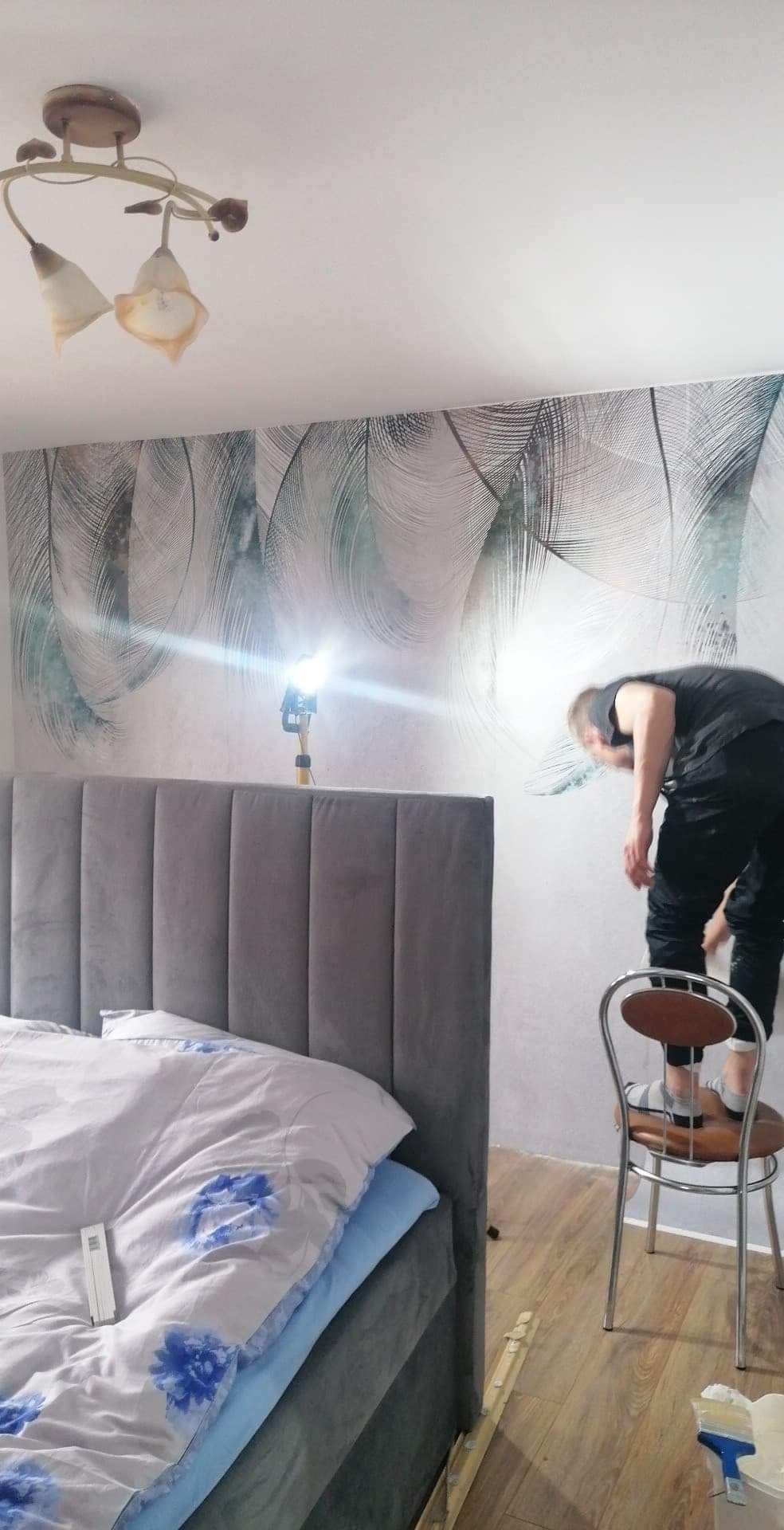 Mężczyzna stojący na krześle, aplikujący wzorzystą tapetę na ścianę w sypialni, oświetloną lampą na statywie, widoczny fragment łóżka z szarym zagłówkiem.