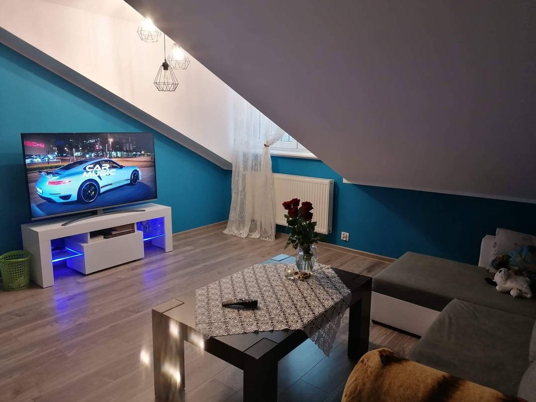 Turkusowa ściana w pokoju na poddaszu, z telewizorem wyświetlającym sportowy samochód, biała szafka RTV z niebieskim podświetleniem LED, stolik kawowy z ażurową serwetą i bukiet czerwonych róż...