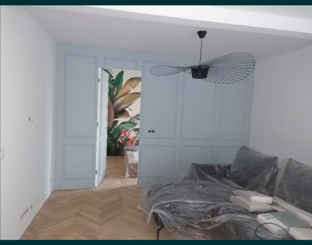 Pokój w trakcie prac wykończeniowych: pomalowane na jasnoniebiesko ściany z dekoracyjnymi panelami, parkiet w jodełkę, designerska lampa wisząca i zabezpieczona folią kanapa.