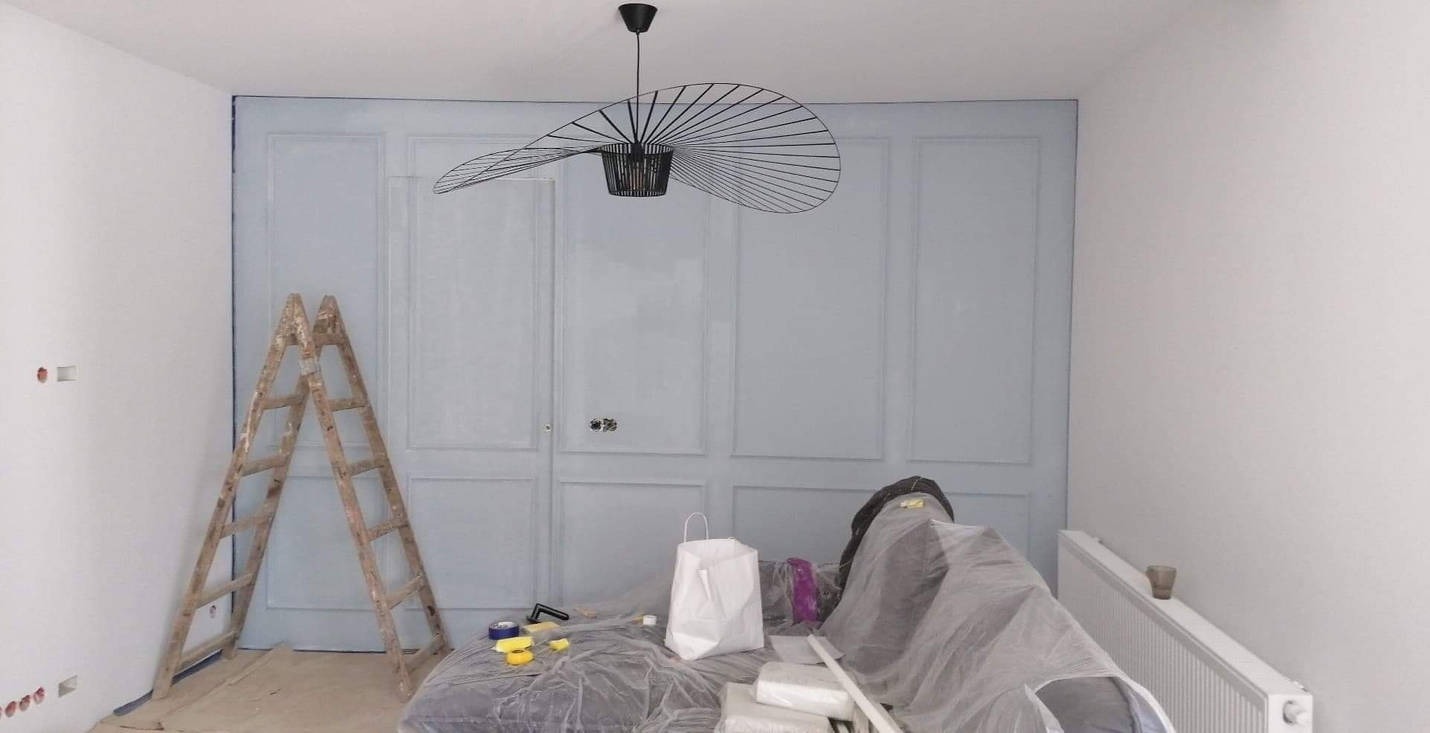 Jasnoszara ściana z panelami w trakcie malowania, widoczna drewniana drabina, designerska lampa wisząca, zabezpieczone meble folią i akcesoria malarskie.