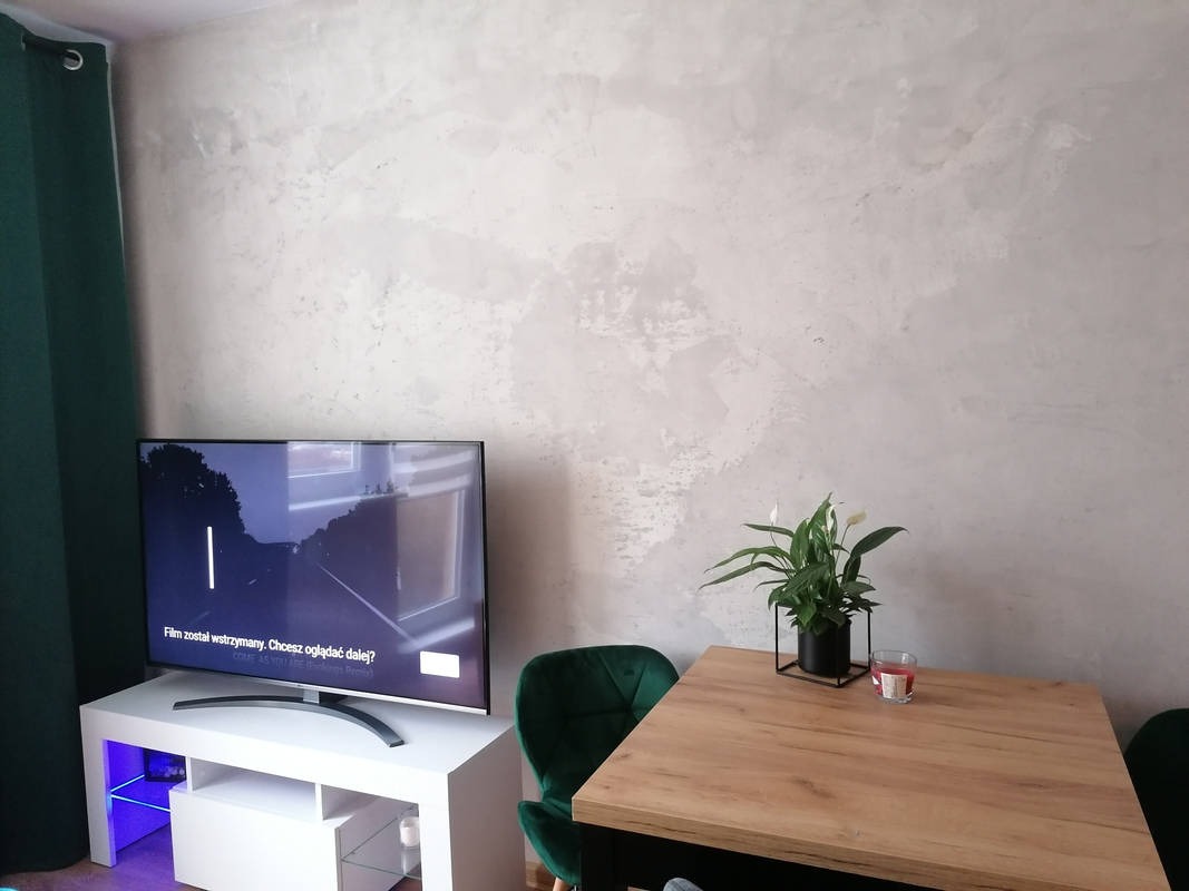Ściana w salonie wykończona dekoracyjnym tynkiem imitującym beton, obok telewizor na białej szafce z podświetleniem LED i drewniany stół z kwiatem w doniczce.