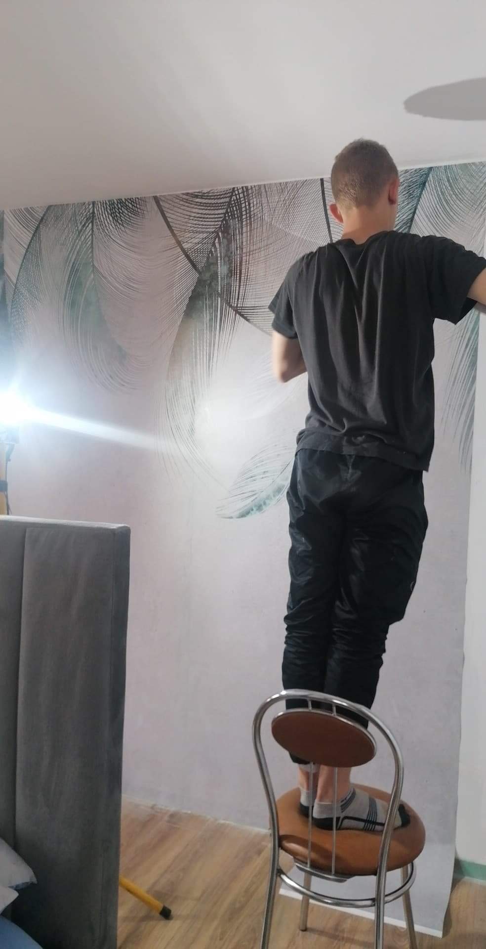 Mężczyzna w czarnej koszulce i spodniach, stojący na krześle, przykleja tapetę z motywem piór na ścianie w pokoju. Widoczny reflektor oświetlający ścianę i fragment szarego zagłówka łóżka.