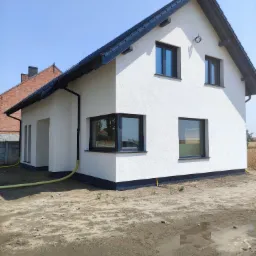 Nowoczesny dom jednorodzinny z białą elewacją i czarną stolarką okienną, widoczny fragment elewacji sąsiedniego domu z czerwonej cegły, teren wokół domu jeszcze niezagospodarowany.