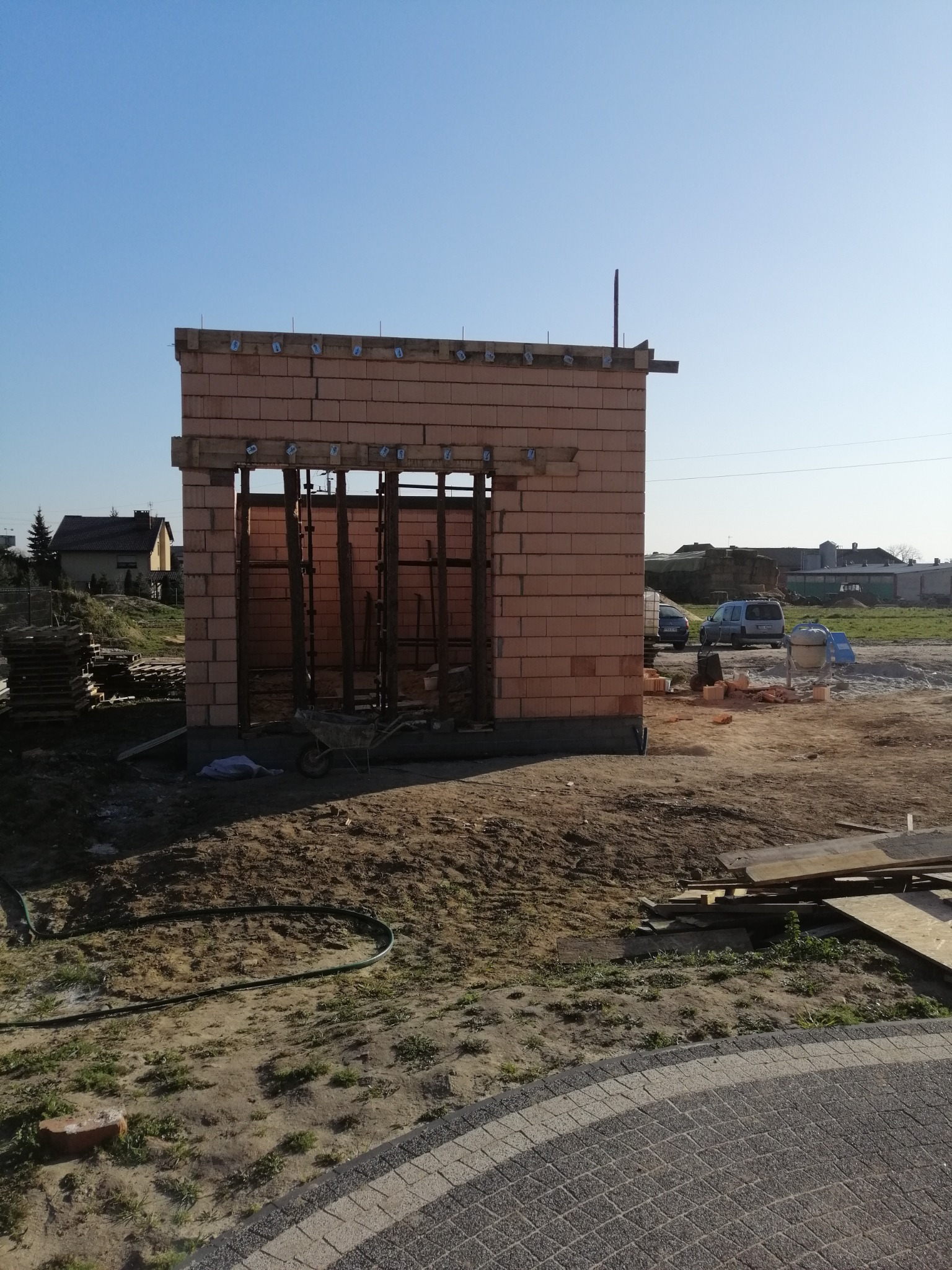 Wznoszona konstrukcja budynku z czerwonej cegły ceramicznej, widoczne drewniane stemple podtrzymujące strop, na zewnątrz plac budowy z gruzem i materiałami budowlanymi.