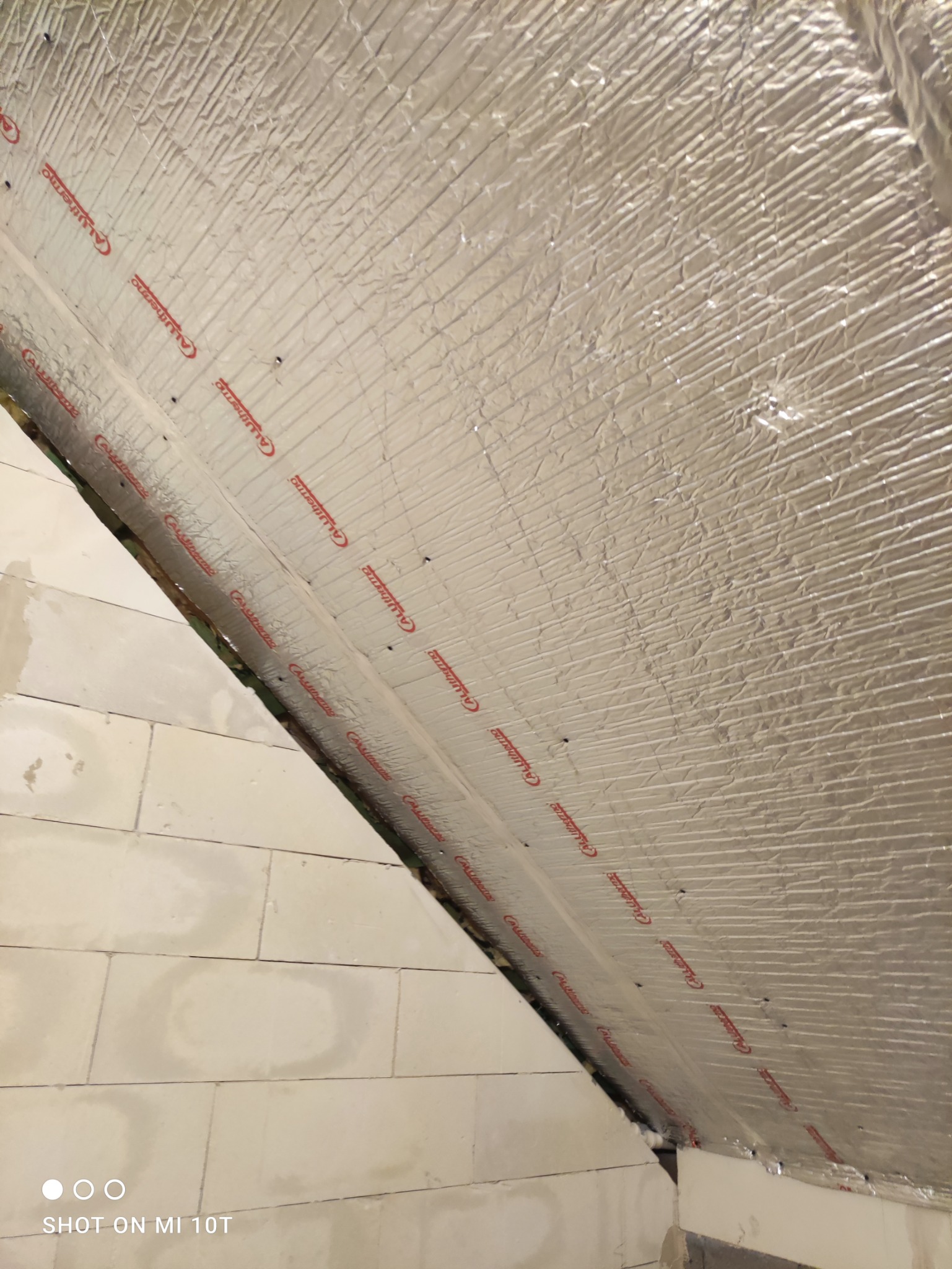 Izolacja termiczna poddasza z folią aluminiową, widoczne bloczki betonowe ściany szczytowej, mocowanie zszywkami, widok z dołu.