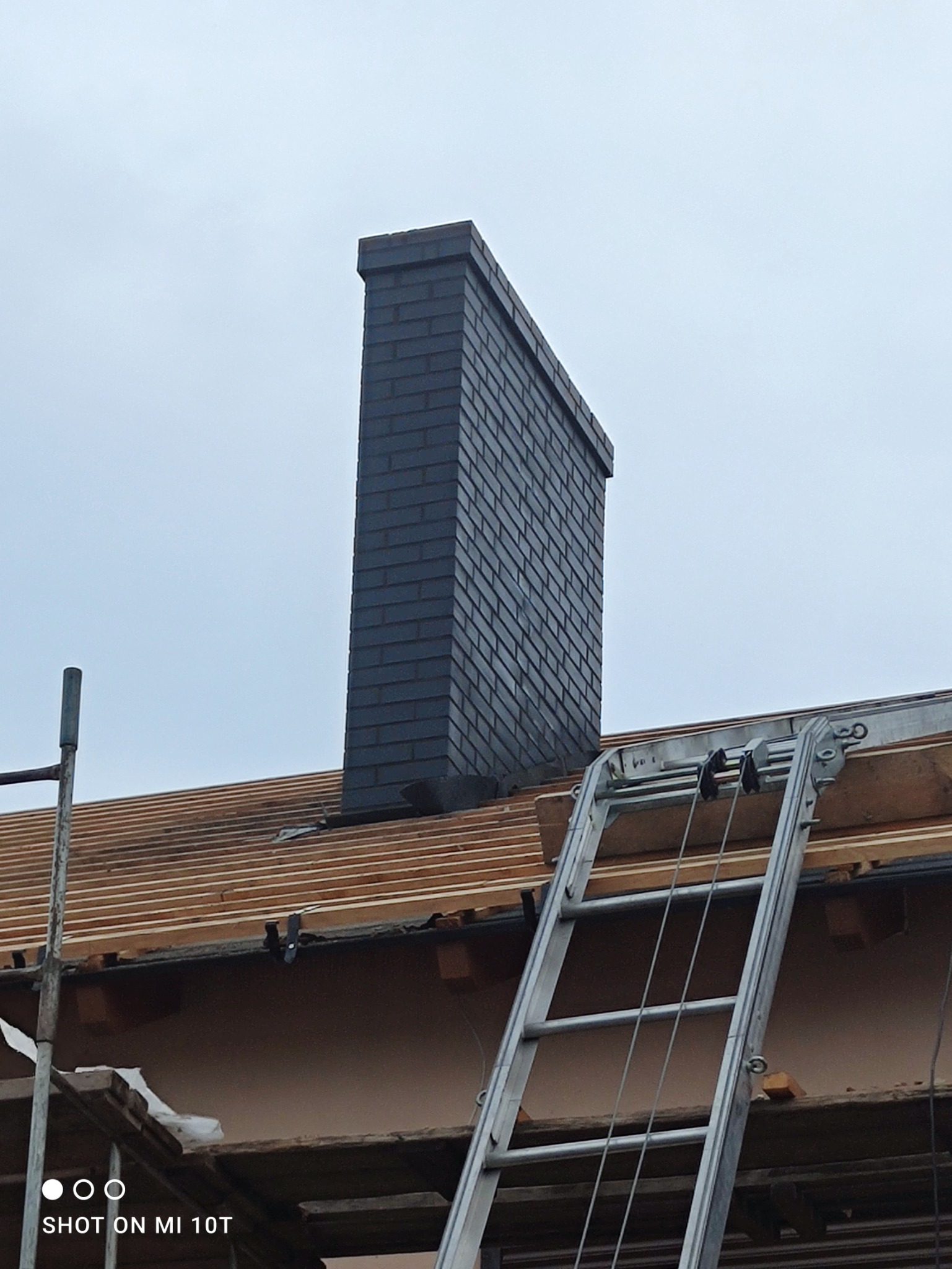 Wznoszony komin z ciemnej cegły na dachu w trakcie budowy, widoczna drabina i rusztowanie.