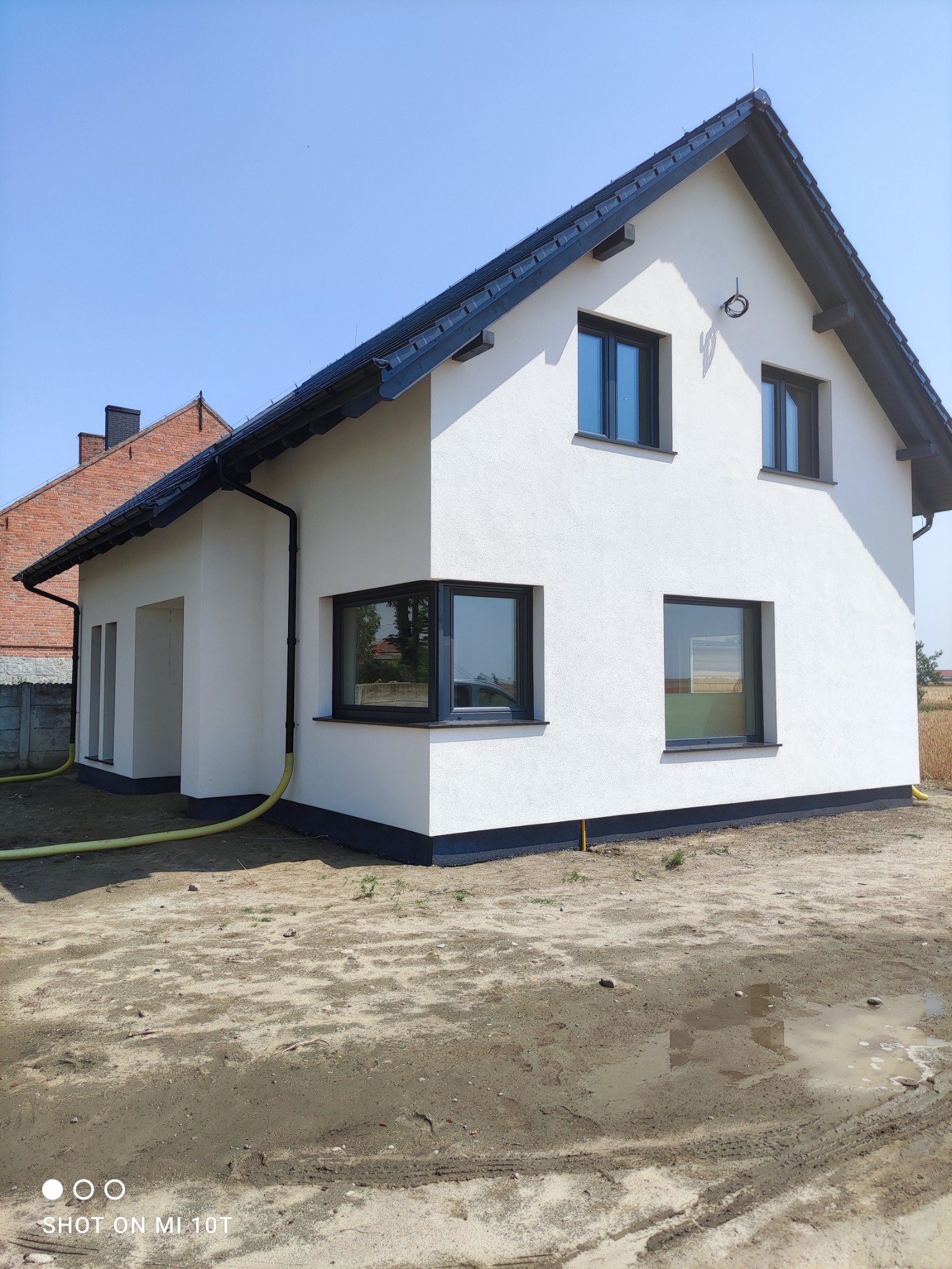 Nowoczesny dom jednorodzinny z białą elewacją i czarną stolarką okienną, widoczny fragment elewacji sąsiedniego domu z czerwonej cegły, teren wokół domu jeszcze niezagospodarowany.